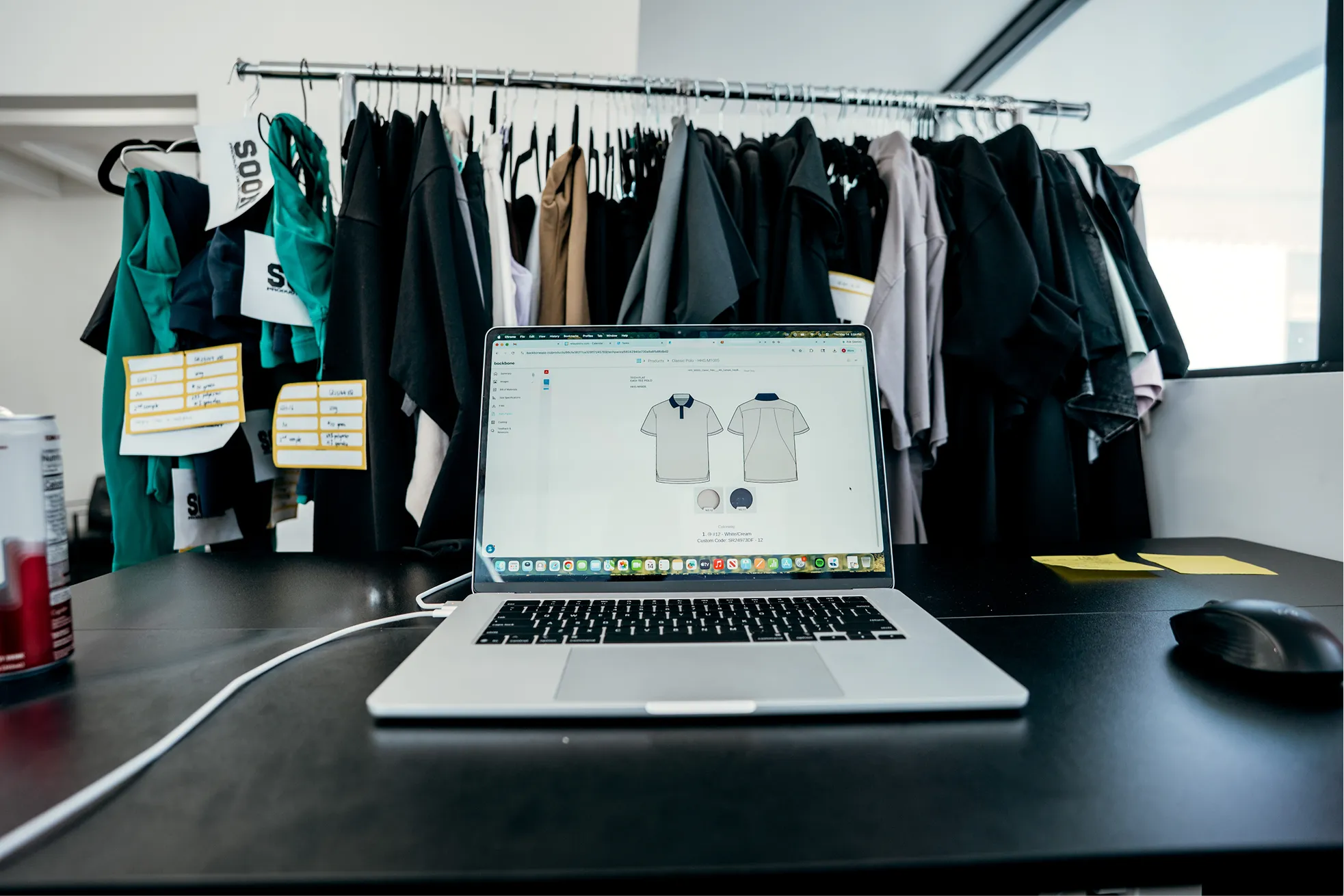 Open laptop on black desk showing polo shirt design, with clothes on hangers and sticky notes in background.