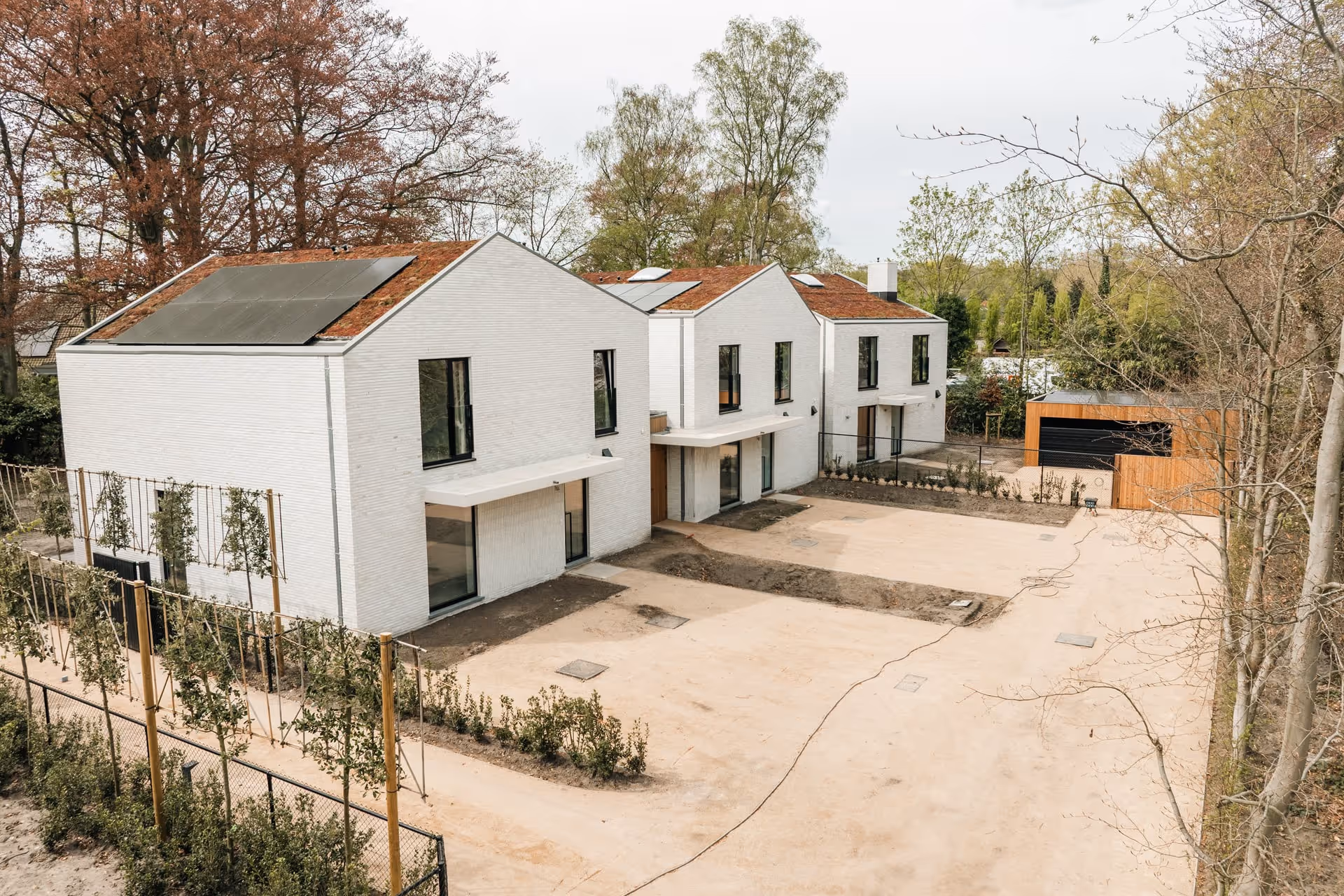 Grondwerken in woonwijk: drie moderne huizen met zonnepanelen, omgeven door bomen.