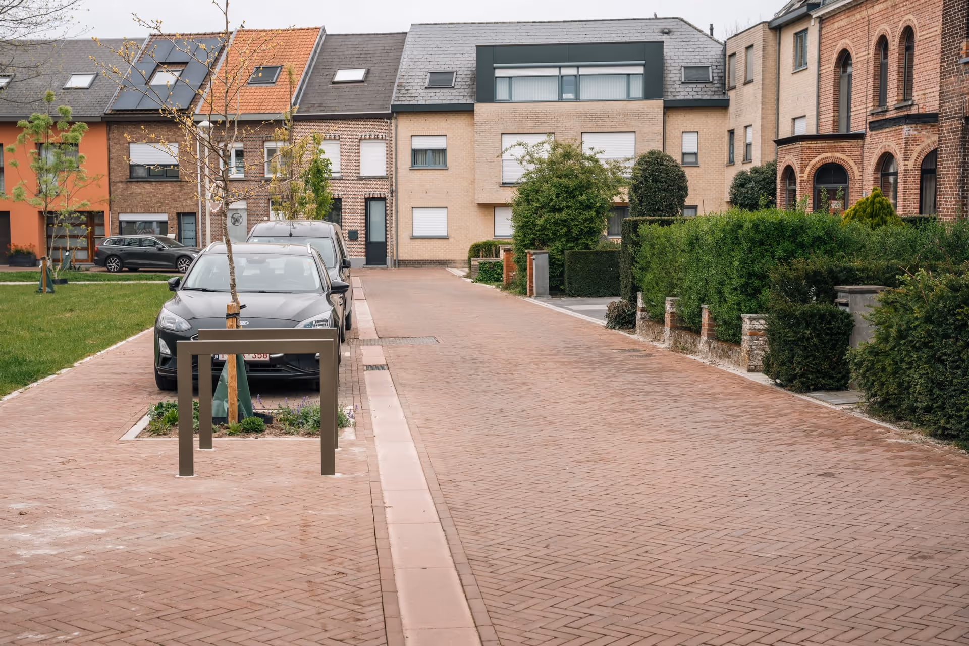 Een woonstraat met geparkeerde auto’s, een met bakstenen geplaveide weg en trottoirs, moderne huizen en groene heggen.