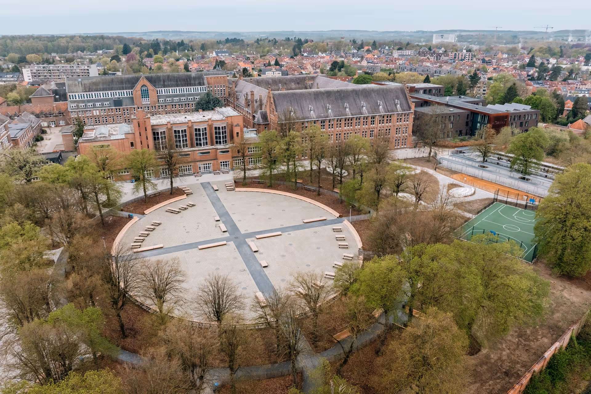 Luchtfoto van een groot schoolterrein met een cirkelvormige, bestrate binnenplaats, bomen rondom, bakstenen gebouwen en een groen sportveld.