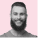 A bearded man smiling against a soft pink background. He wears a casual t-shirt, projecting a friendly and approachable demeanor.