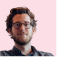 A man with curly hair and glasses smiles slightly against a light pink background. He wears a dark shirt, conveying a relaxed, approachable mood.
