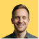 A smiling person with short hair is in front of a solid yellow background, conveying a friendly and positive demeanor.