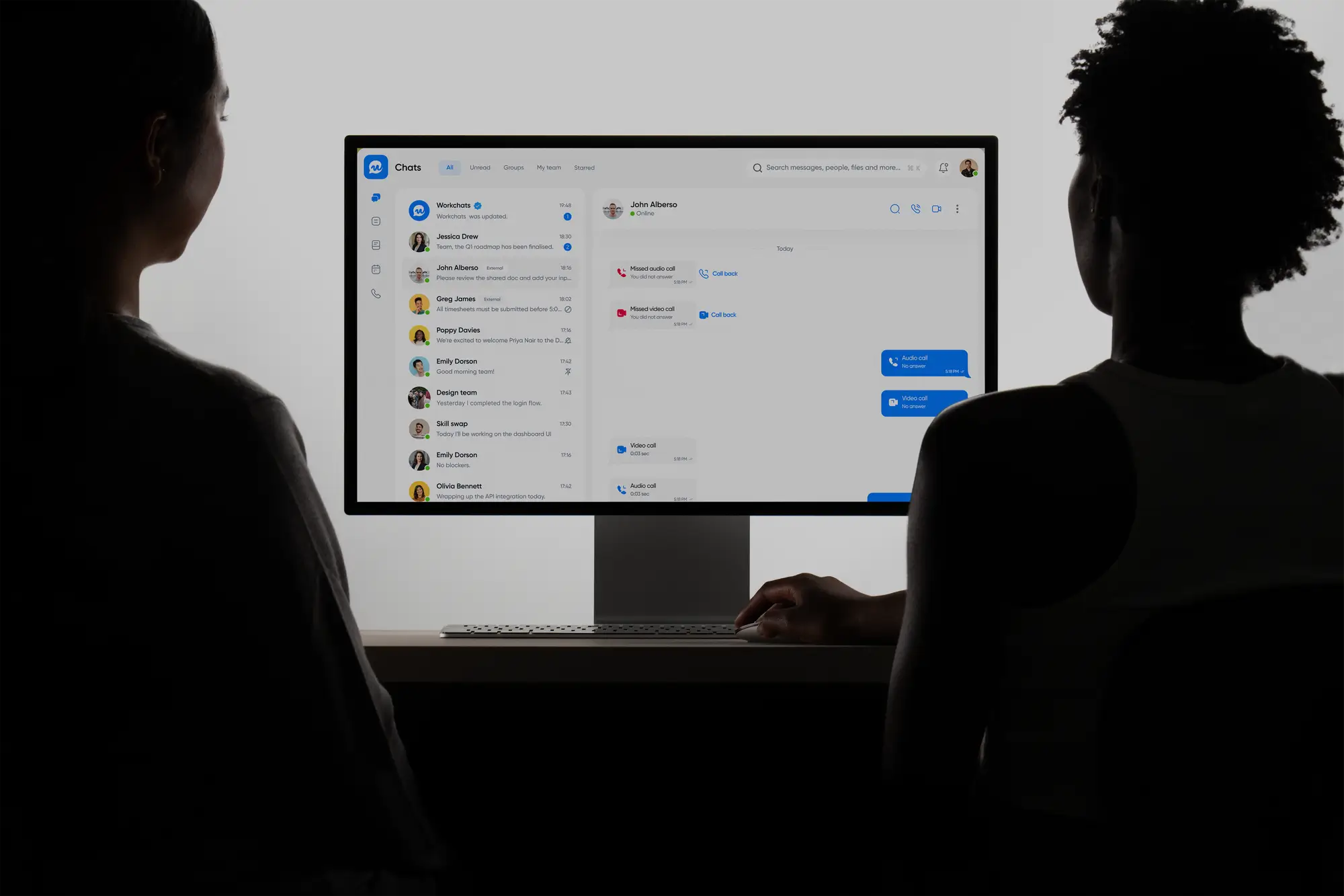 Two individuals sit in front of a computer screen displaying a messaging app. The room is dimly lit, highlighting the illuminated screen's interface.