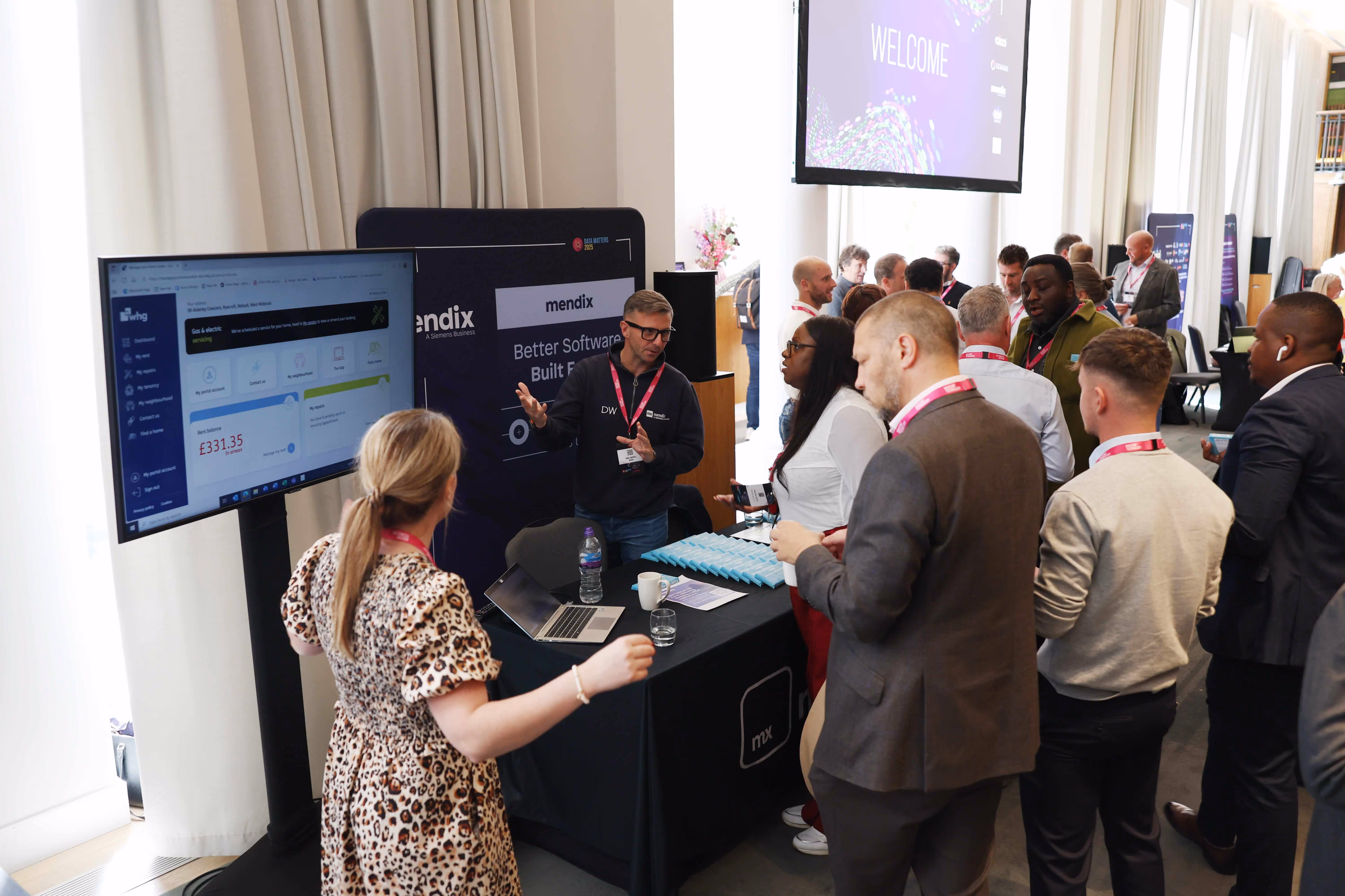 People gathered around a Mendix booth at a conference, with a man explaining software on a laptop and large screen.