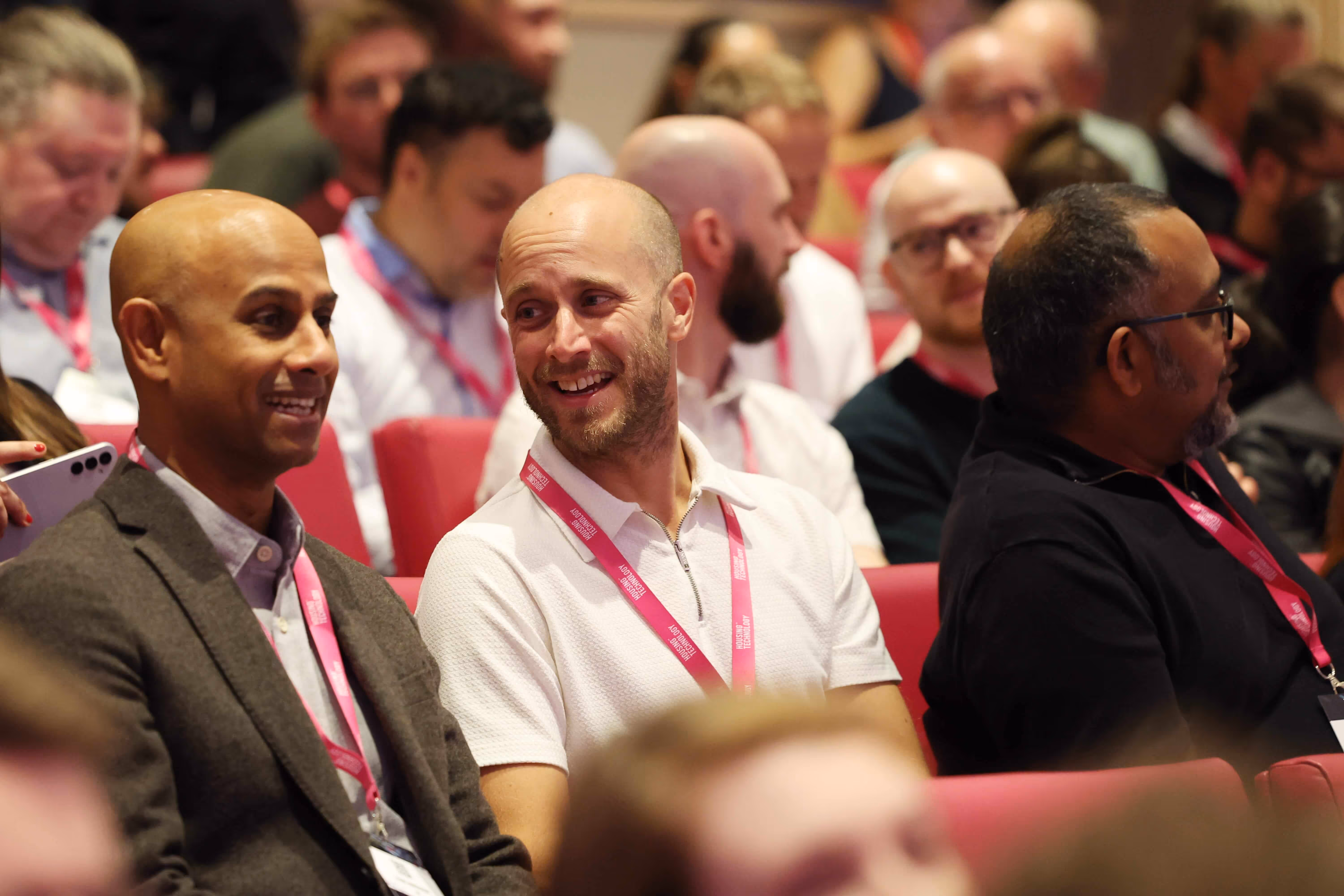 Three men wearing conference badges sitting in a red auditorium, two of them smiling and talking.