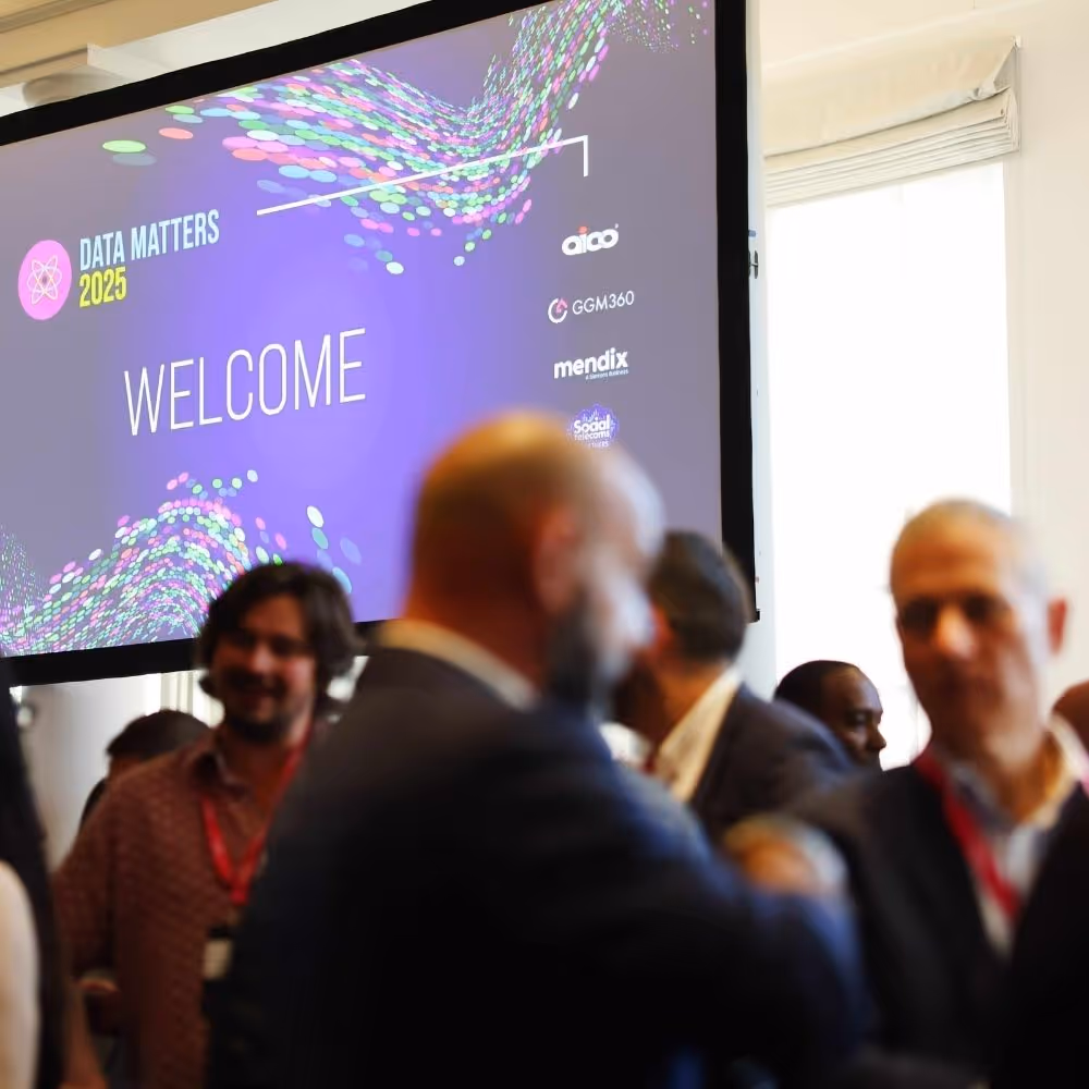 Conference room with attendees networking and a large screen displaying 'Data Matters 2025 WELCOME' and several sponsor logos.