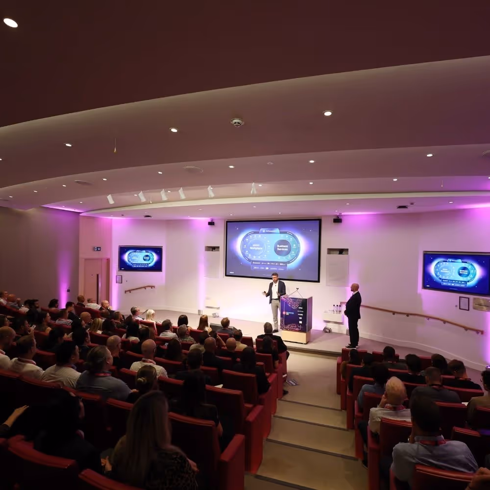 Speaker presenting in a conference room with audience seated in red chairs and digital screens displaying a business services diagram.