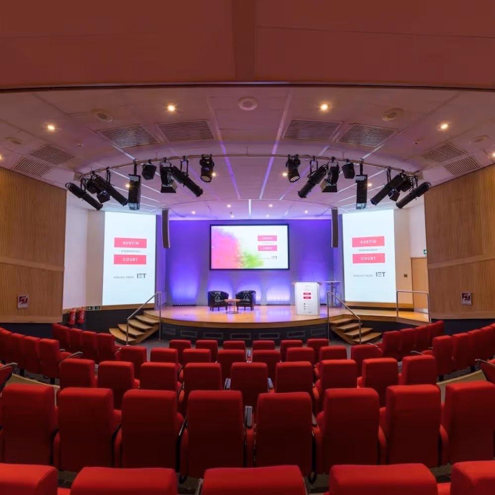 Empty auditorium with red seats facing a stage with two chairs, a small lectern, and projection screens displaying 'Austin Court' and IET logos.