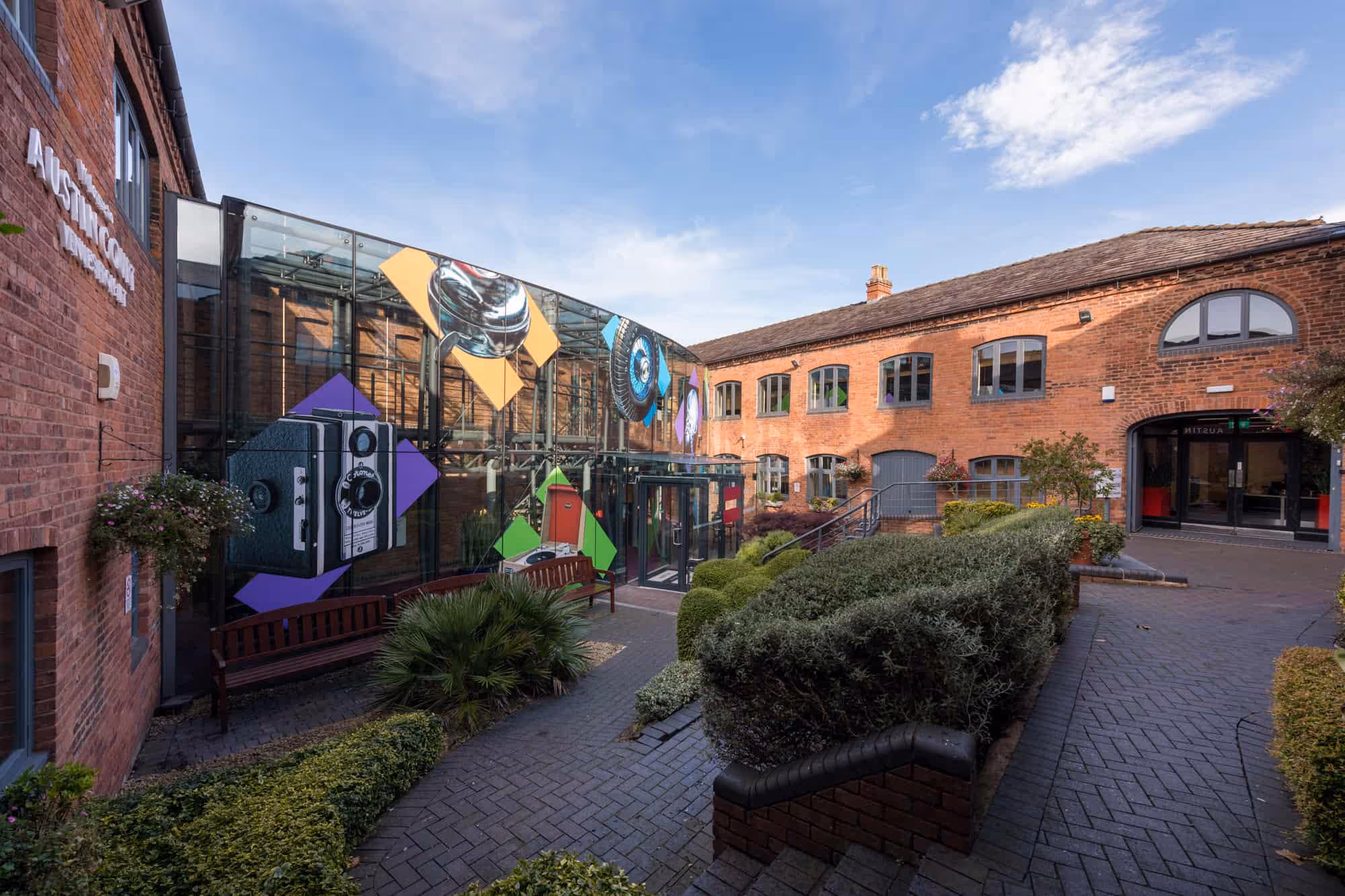 Image of IET Austin Court with view of the courtyard with brick buildings, glass facade adorned with colorful camera graphics, benches, and manicured shrubbery under a blue sky.