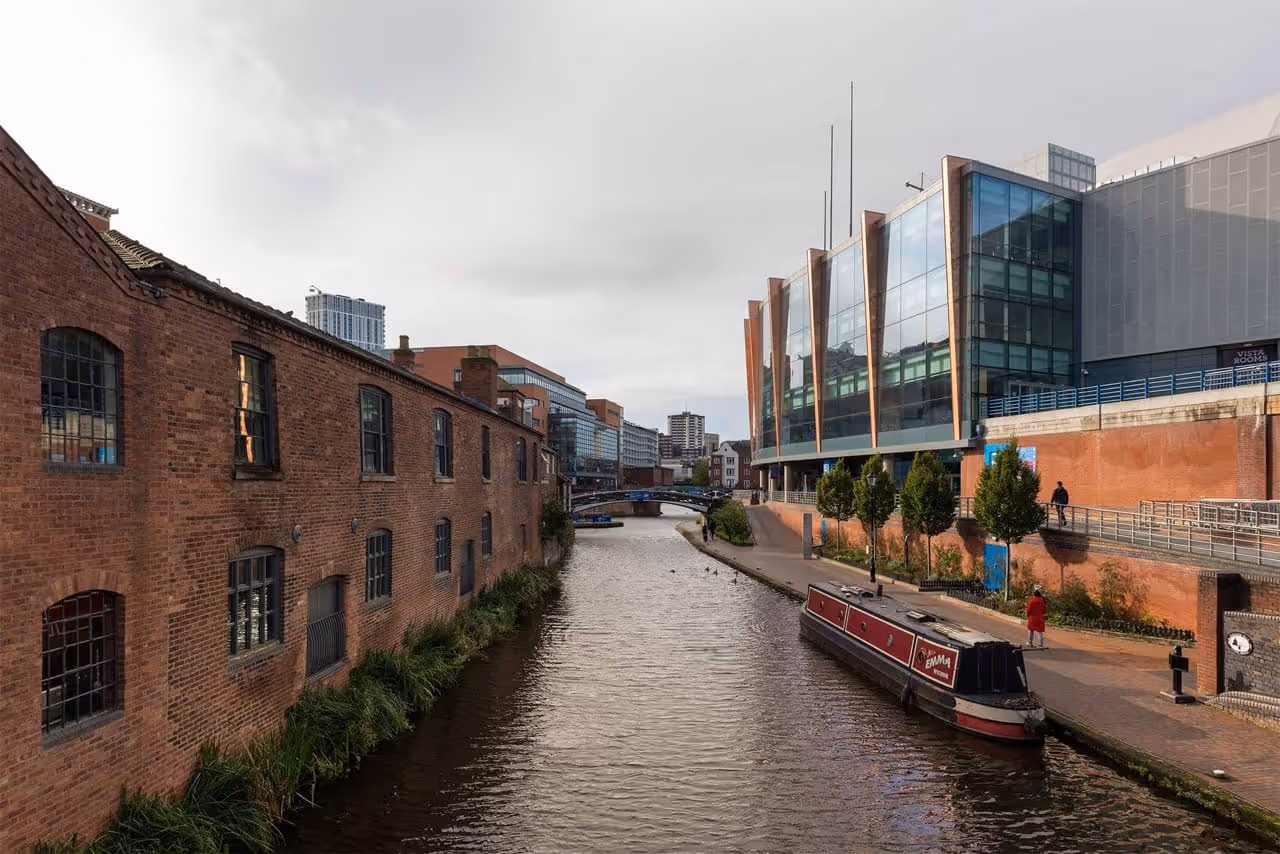 View of Birmingham canal with a narrowboat docked alongside a brick walkway, flanked by an old brick building on the left and a modern glass building on the right.