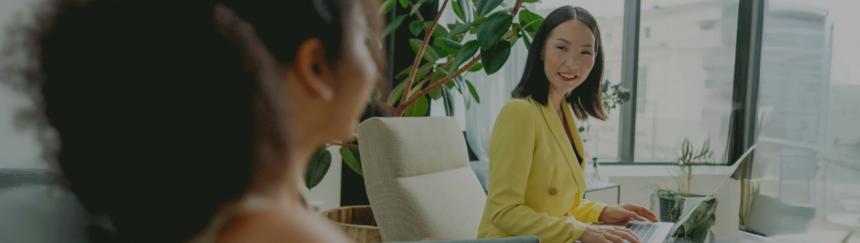 Woman in yellow blazer smiling and using a laptop, with another person blurred in the foreground in a modern office setting.