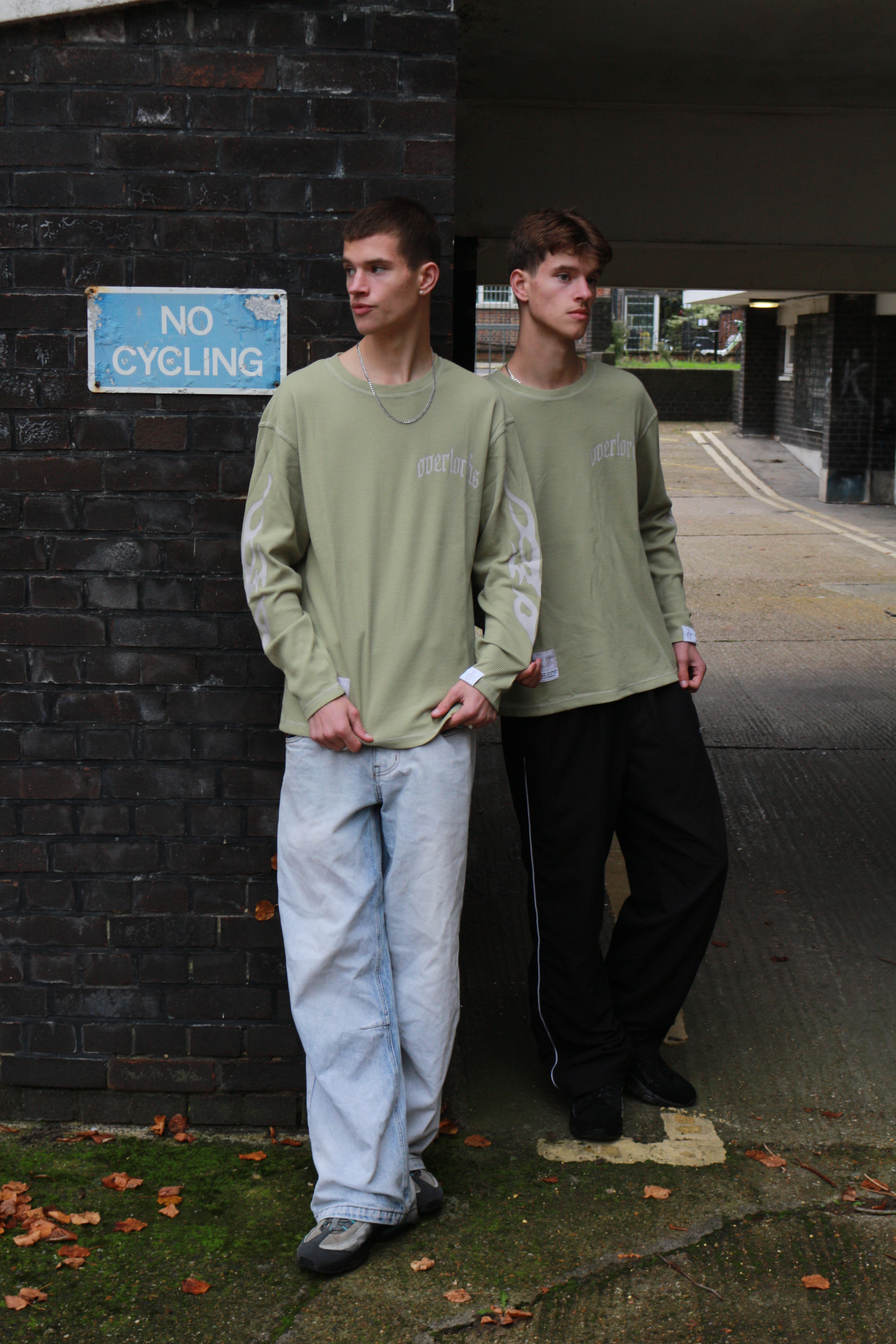Two young men wearing matching olive green long-sleeve shirts stand against a brick wall near a 'No Cycling' sign in an urban alleyway.
