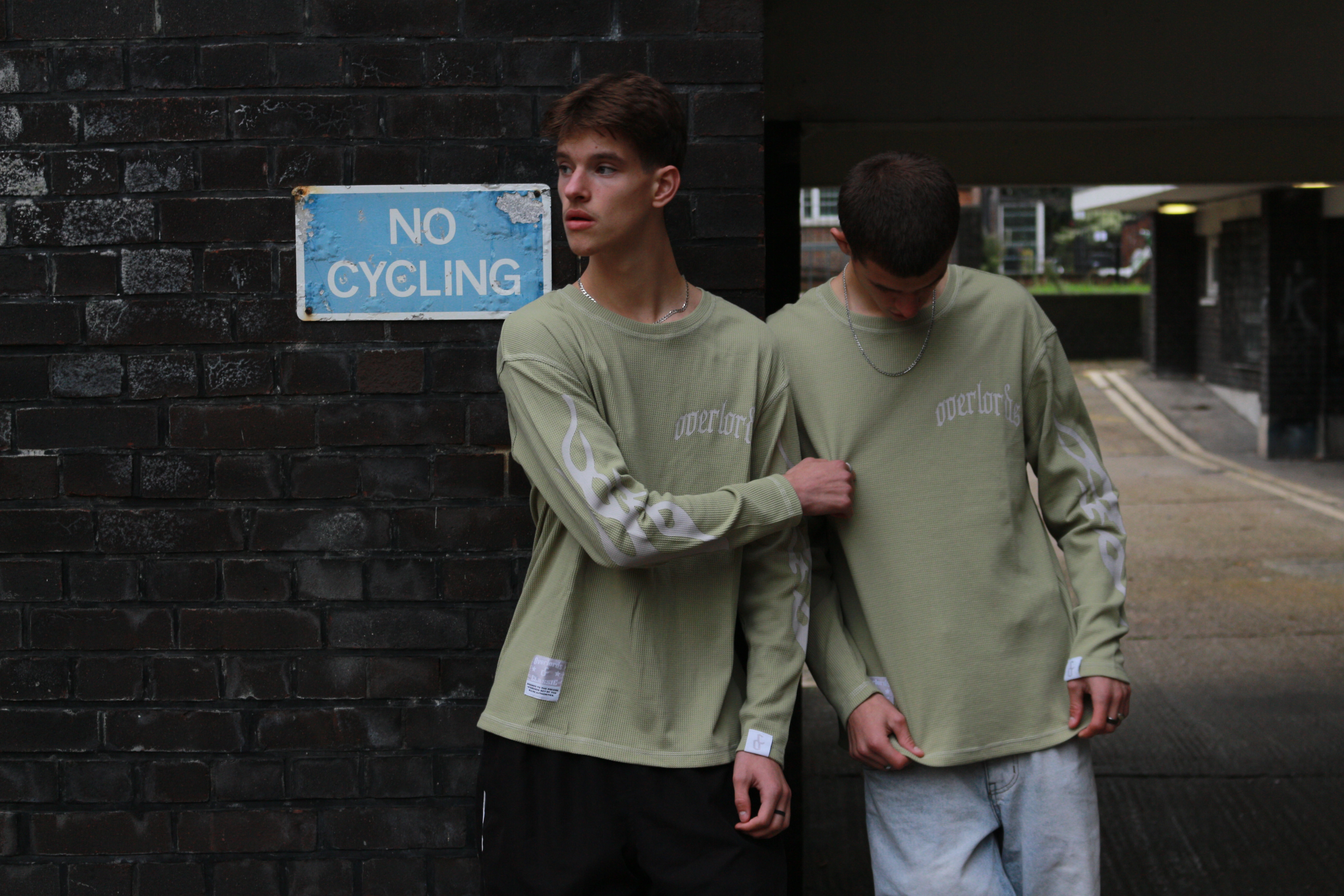 Two young men wearing matching light green long-sleeve shirts stand near a black brick wall with a faded blue 'NO CYCLING' sign.