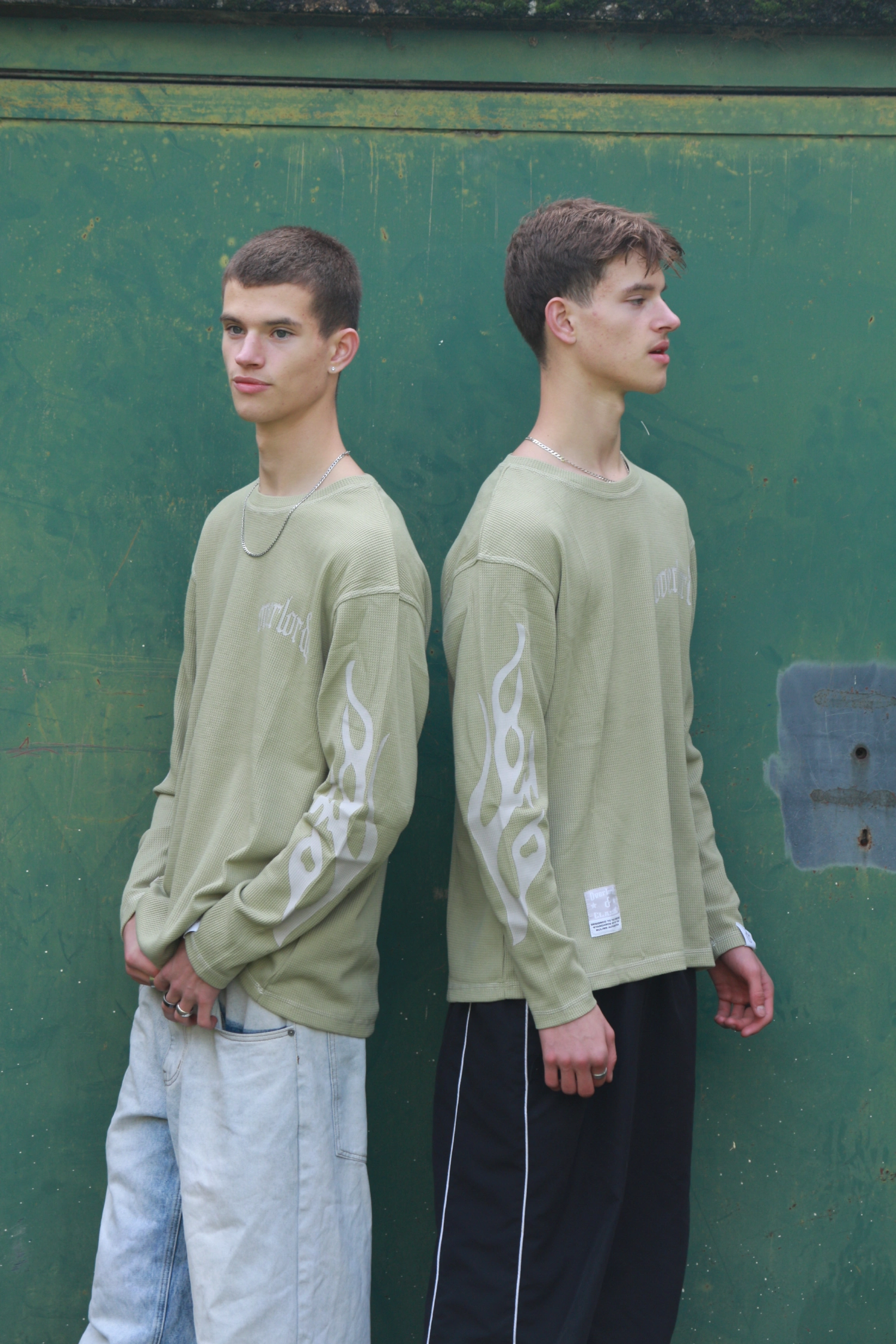 Two young men standing back to back against a green wall, both wearing light green long-sleeve shirts with white flame designs on the sleeves.