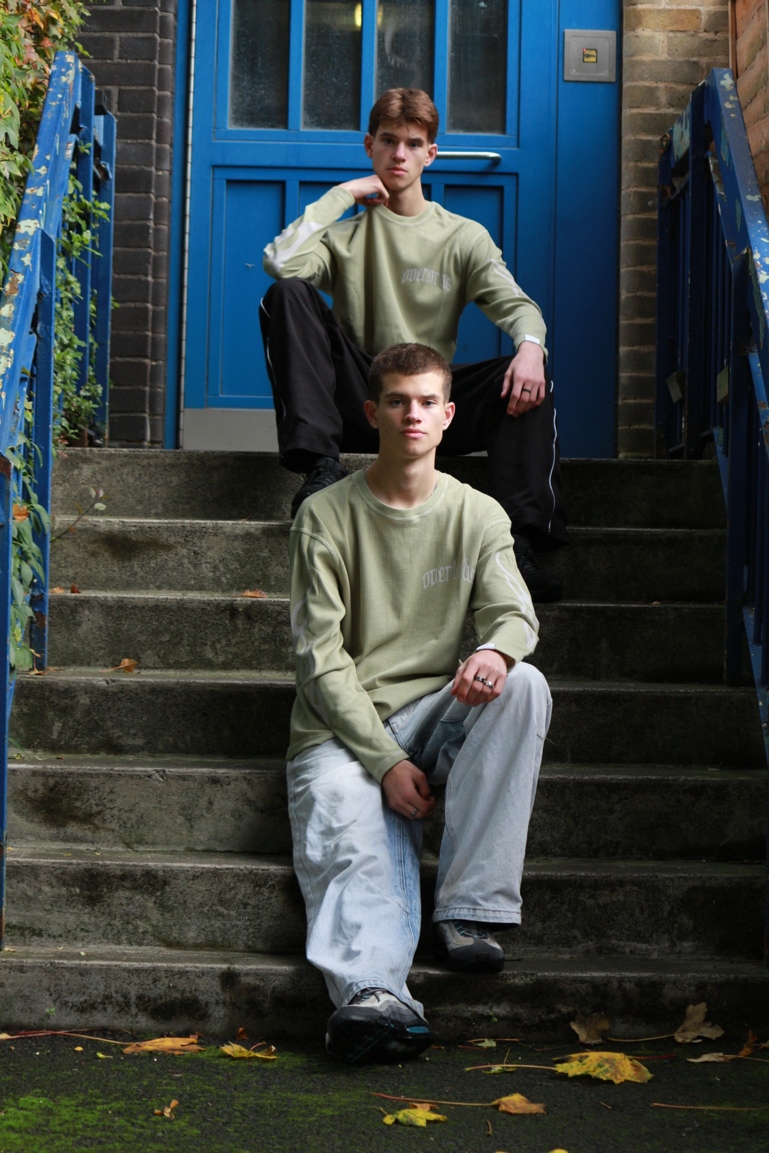 Two young men sitting on concrete steps in front of a blue door, both wearing light green long-sleeve shirts.