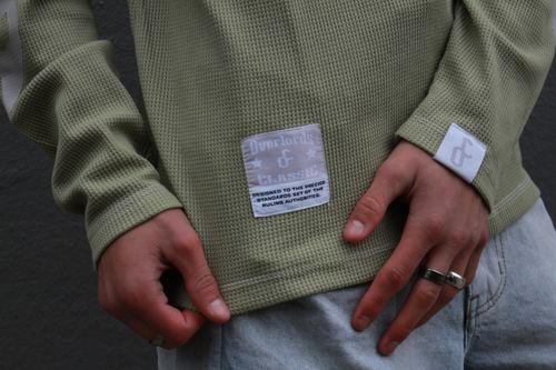 Close-up of a person's hands wearing an olive green waffle-knit long-sleeve shirt with a label that reads 'Overlords & Classic' and light blue jeans.