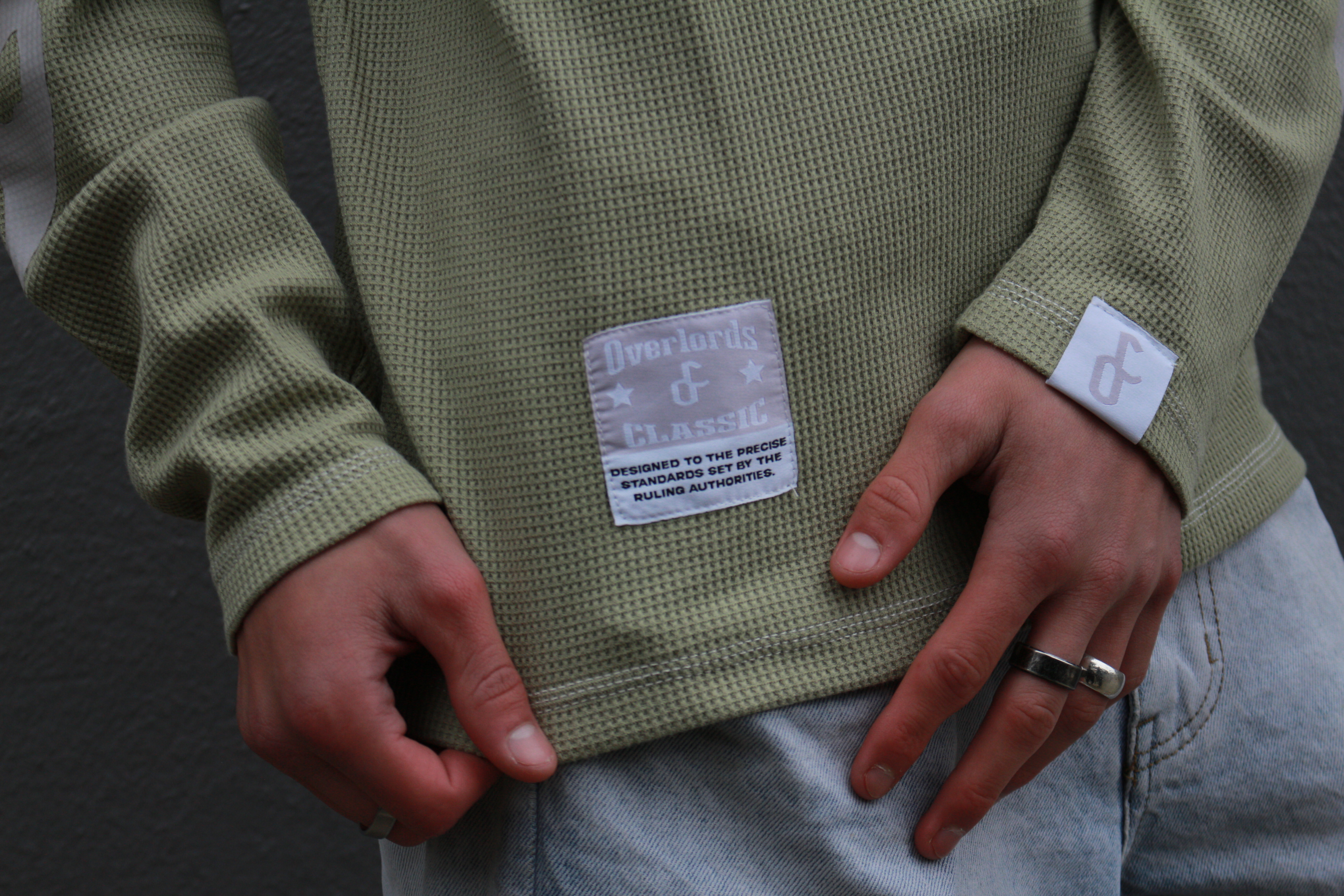 Close-up of a person's hands wearing an olive green waffle-knit long-sleeve shirt with a label that reads 'Overlords & Classic' and light blue jeans.