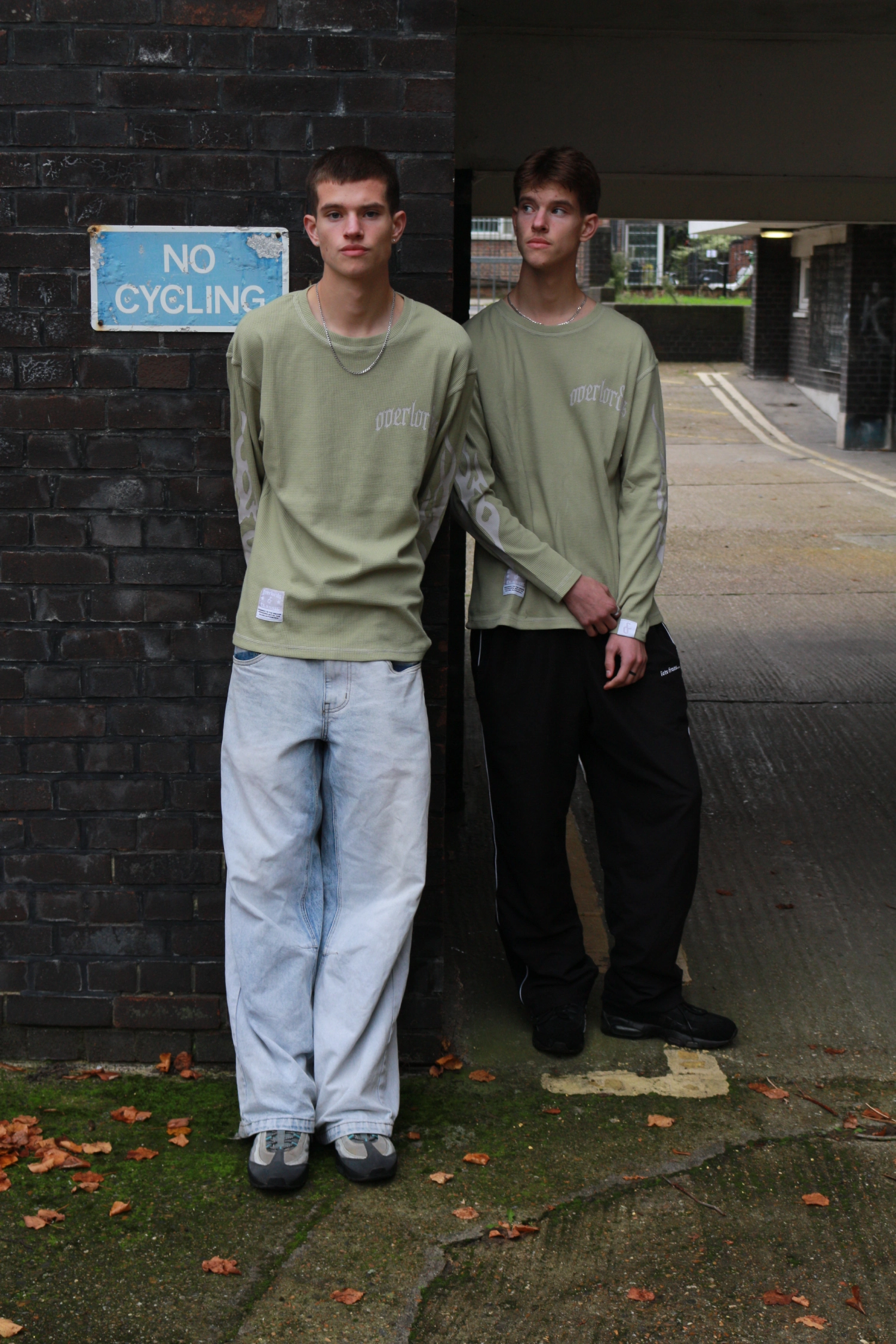 Two young men dressed similarly in light green long-sleeve shirts standing against a brick wall with a 'No Cycling' sign.