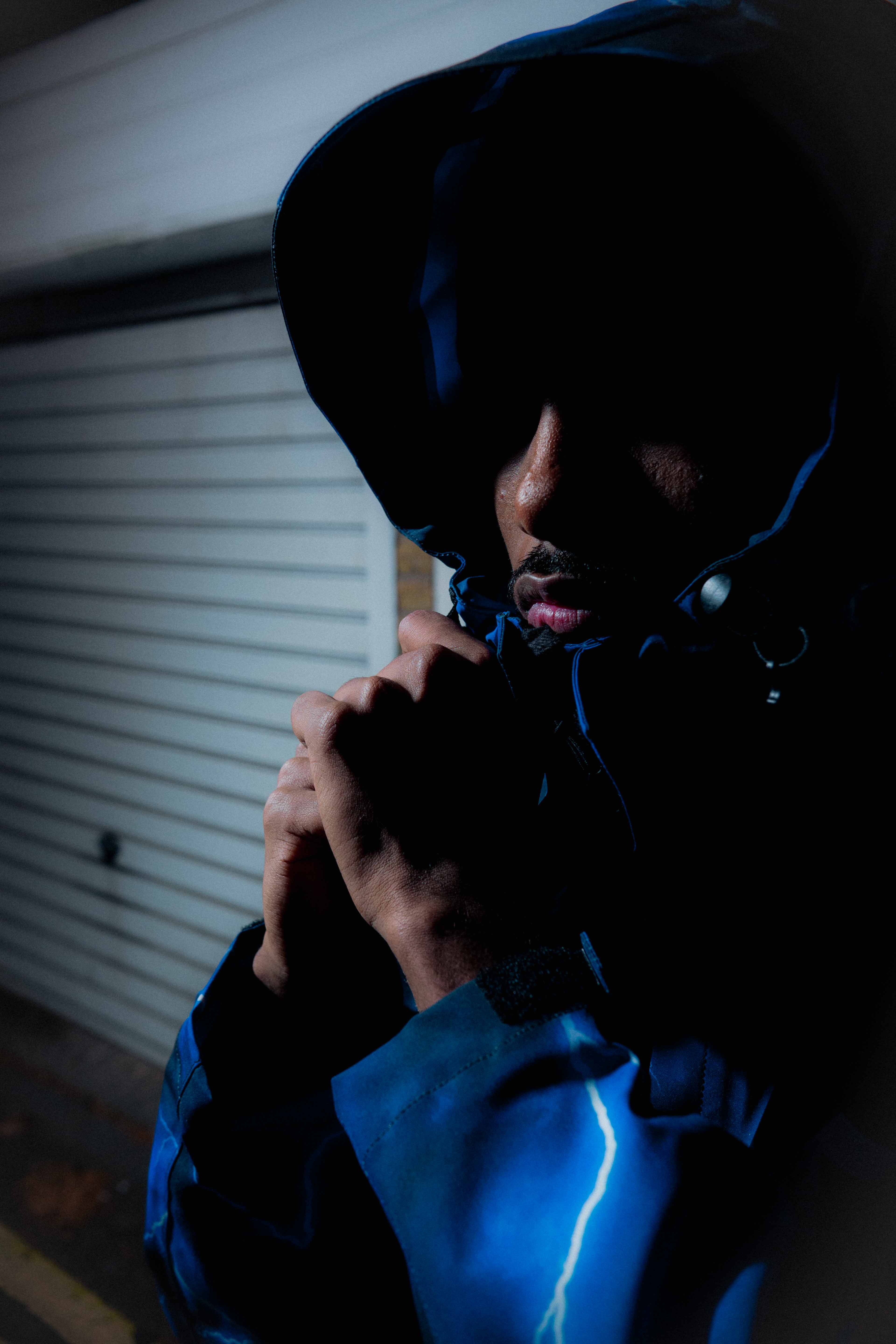 Man in a blue jacket with a hood pulled tight, partially illuminated against a dim background.