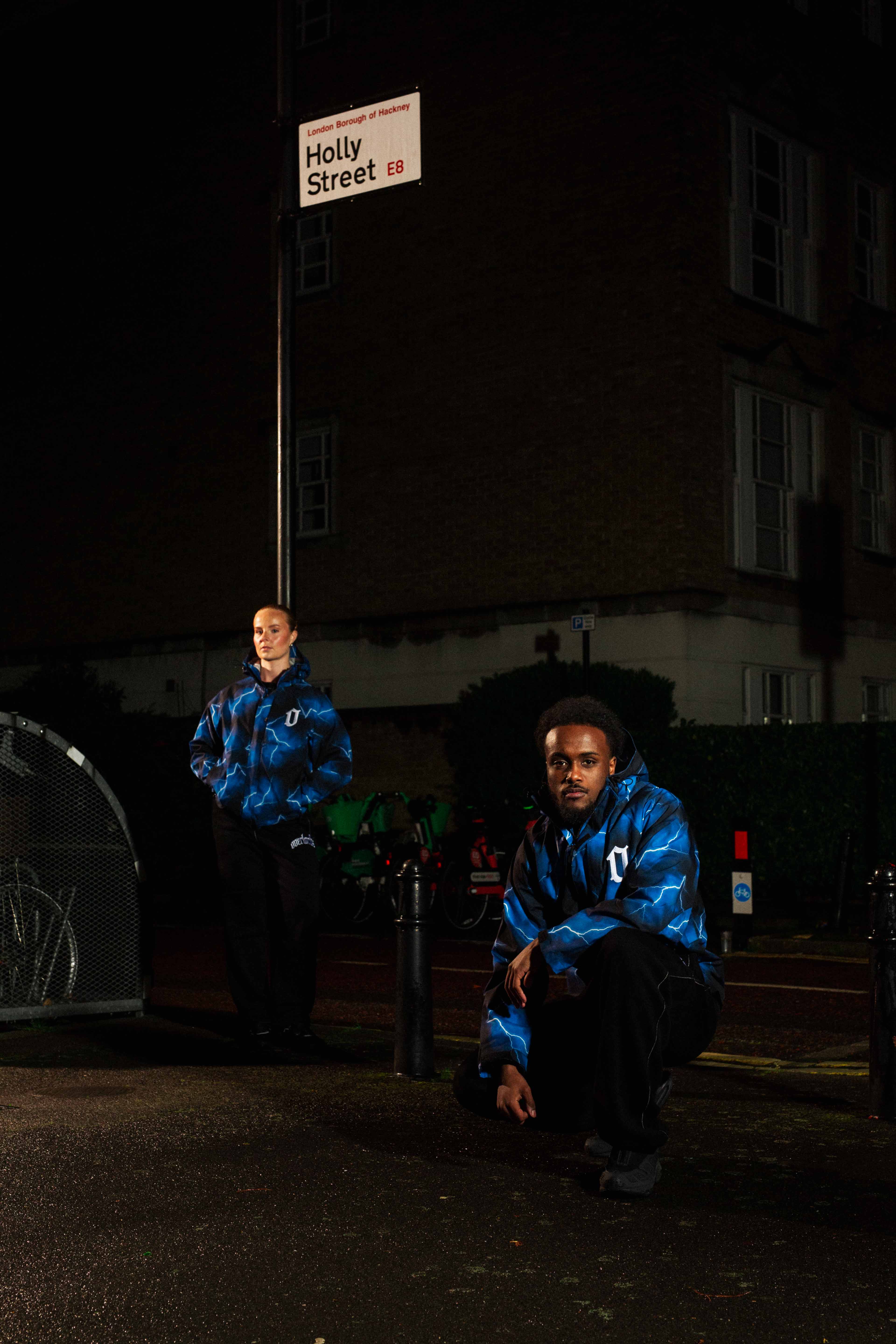 Two people wearing matching blue lightning-patterned jackets posing on a dimly lit street under a Holly Street sign in London.