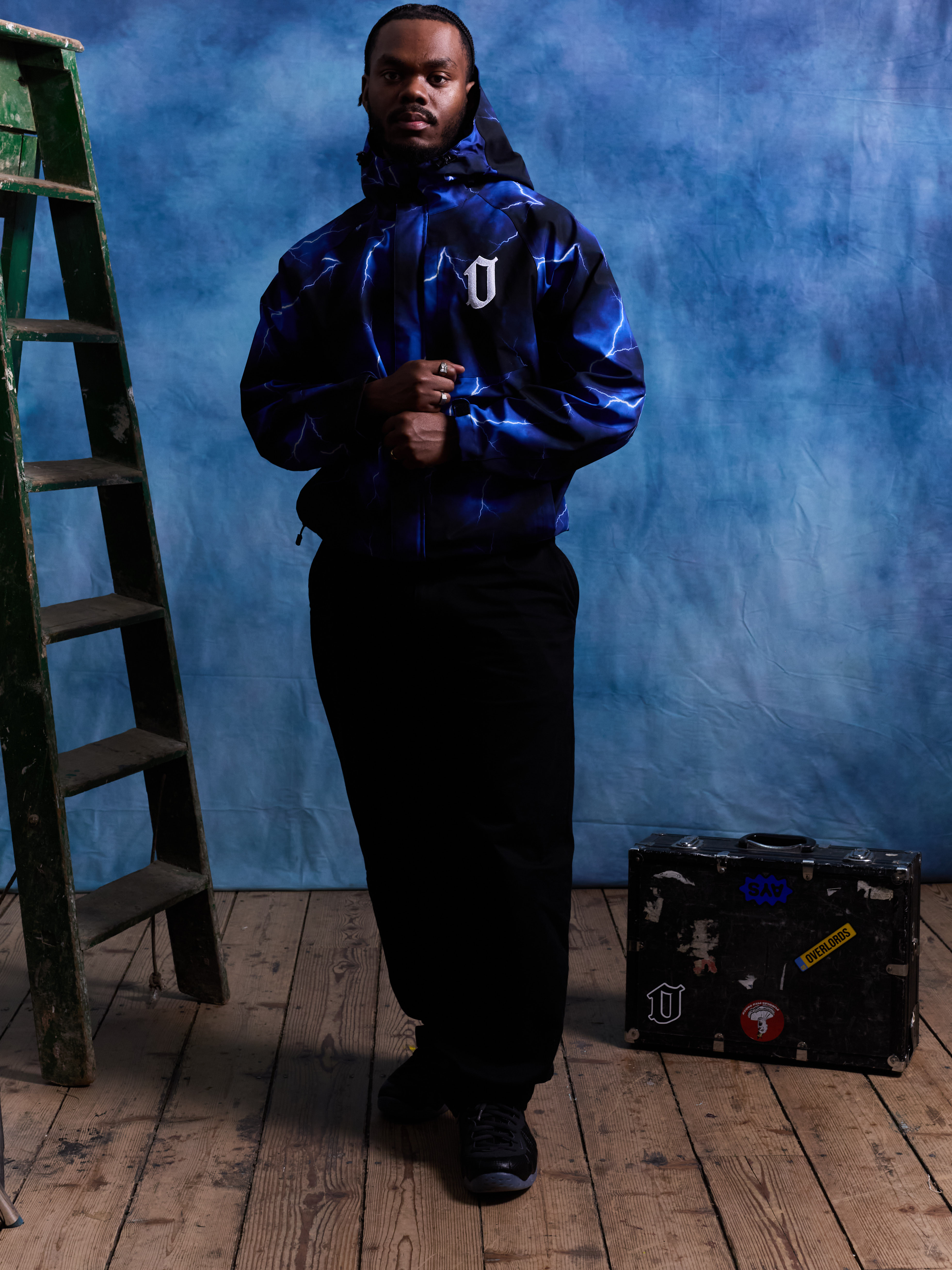 Man in a blue lightning-patterned hooded jacket standing with clasped hands next to a green ladder and a black suitcase with stickers on a wooden floor.