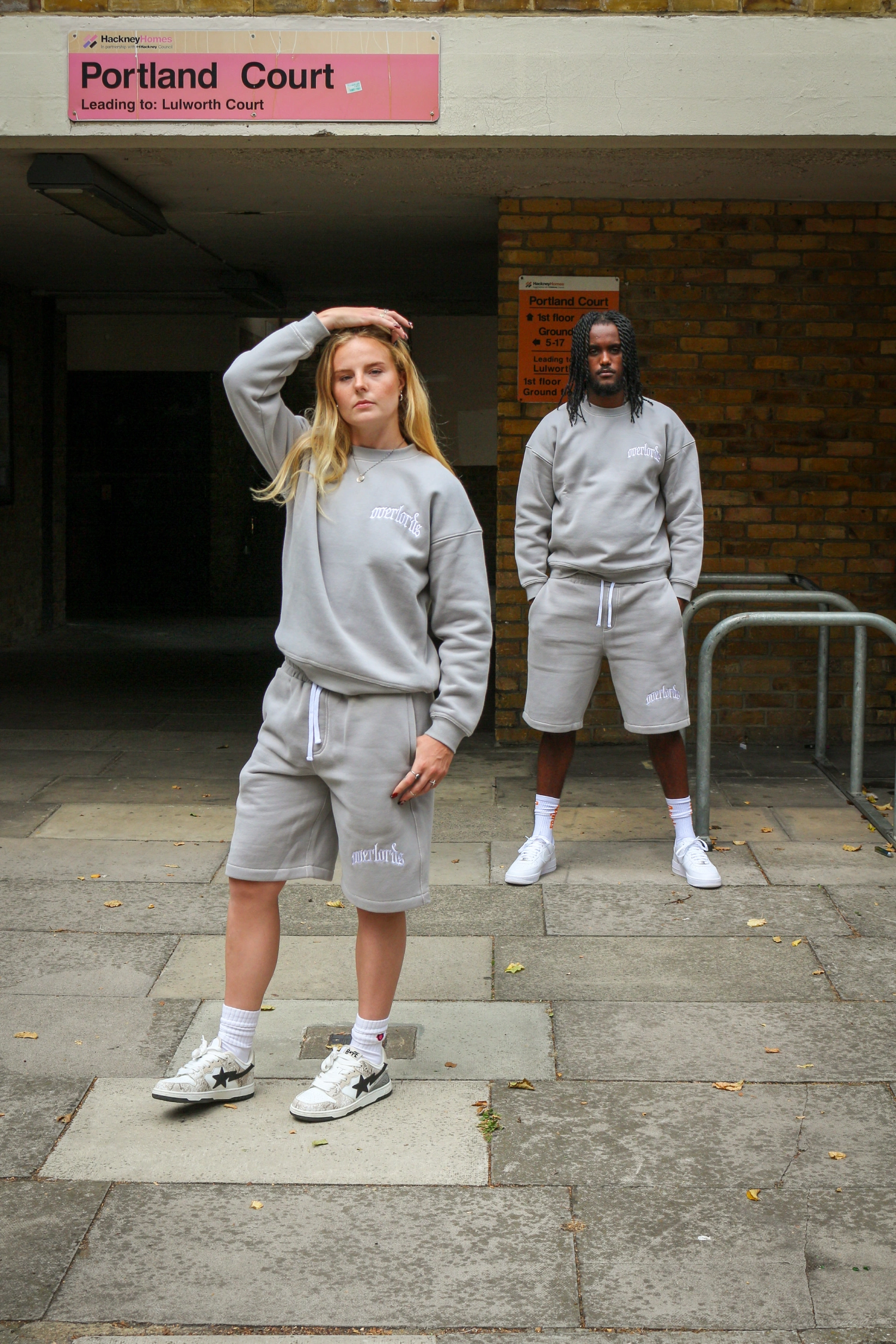 Two models wearing matching gray sweatshirts and shorts standing on a paved walkway in front of a Portland Court sign.