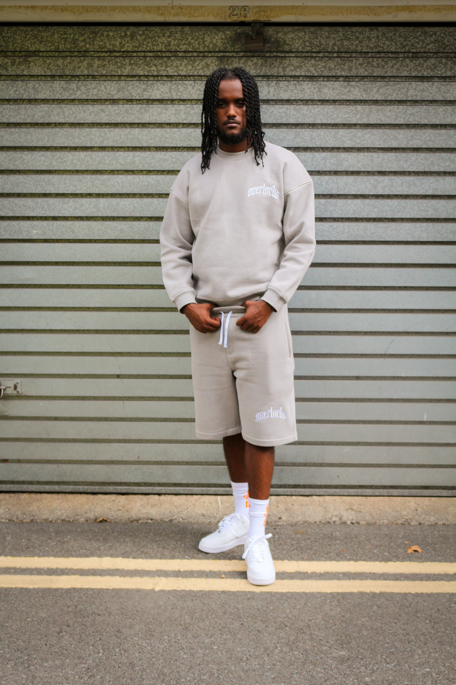Man with braided hair standing in front of a gray garage door wearing a beige sweatshirt and matching shorts with white sneakers and socks.