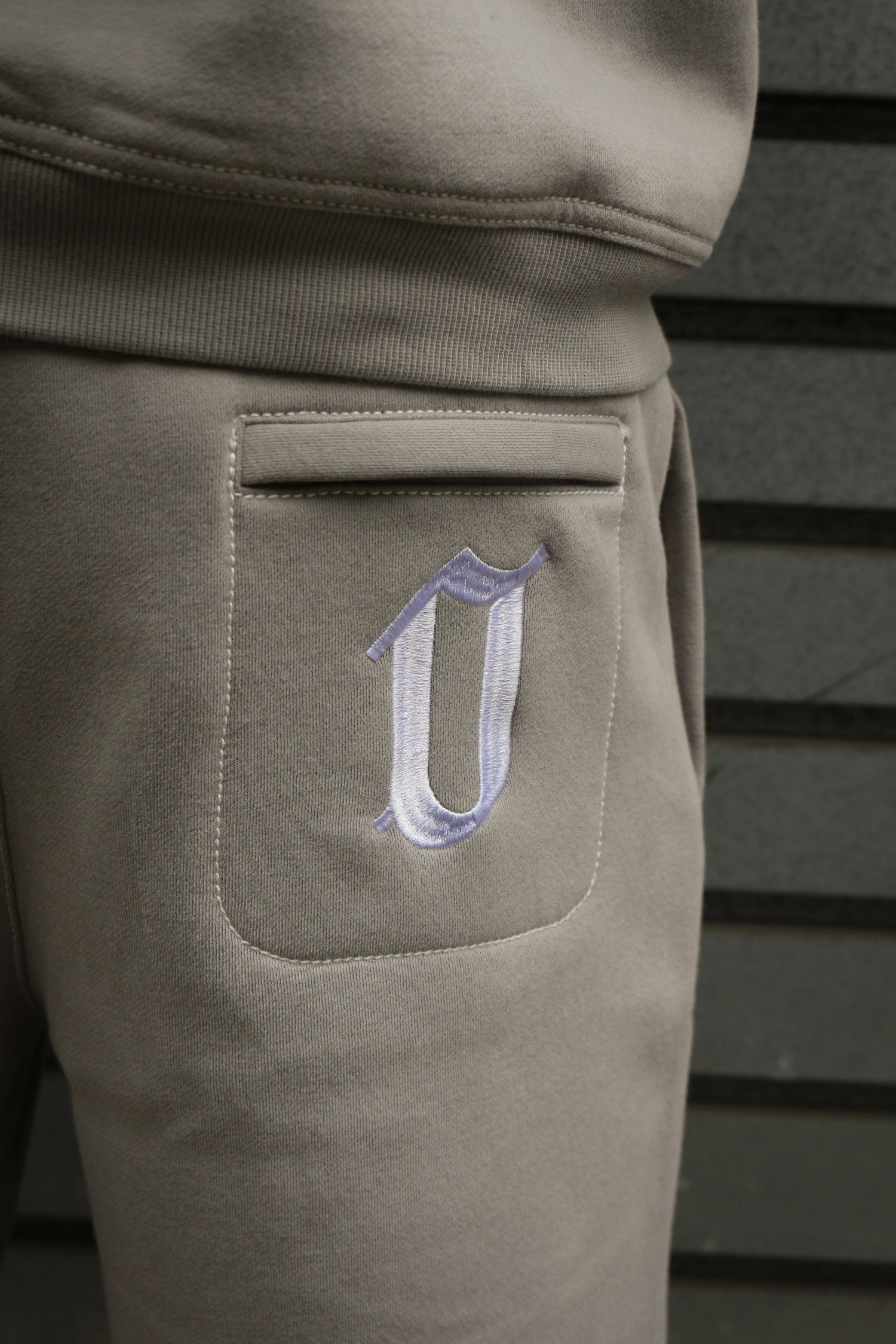 Close-up of beige sweatpants pocket with a white and light purple embroidered letter U.