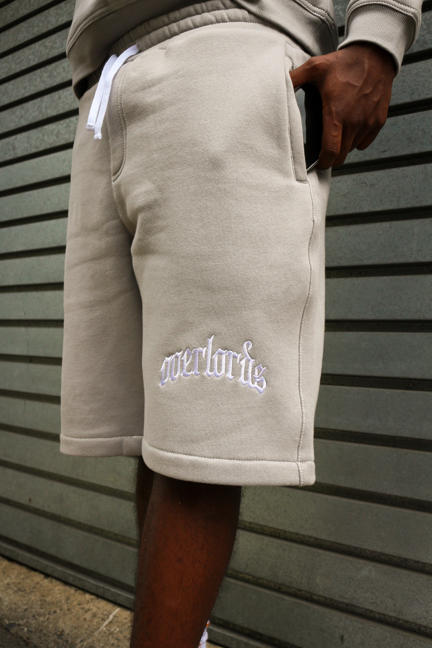 Person wearing beige shorts with white embroidered text 'overlorius' near the hem, with a hand holding a phone in the pocket.