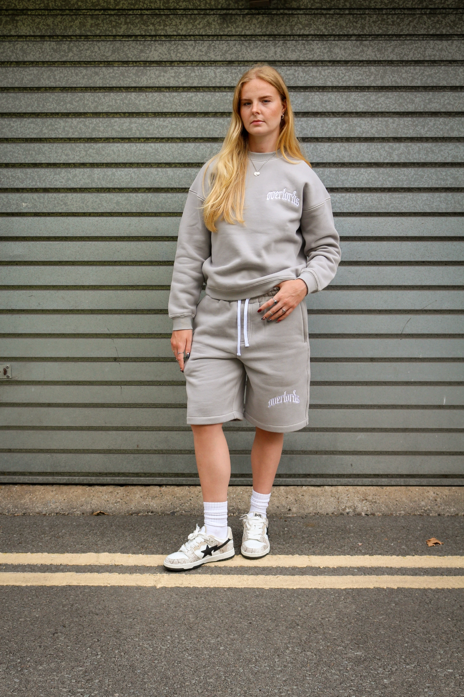 Woman with long blonde hair wearing light gray sweatshirt and shorts standing on a street with gray metal shutter background.