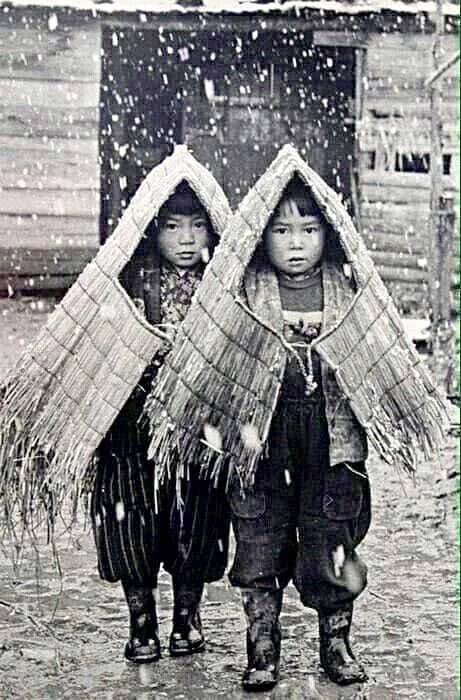 Two children standing in the snow wearing traditional Japanese rain gear made of straw.