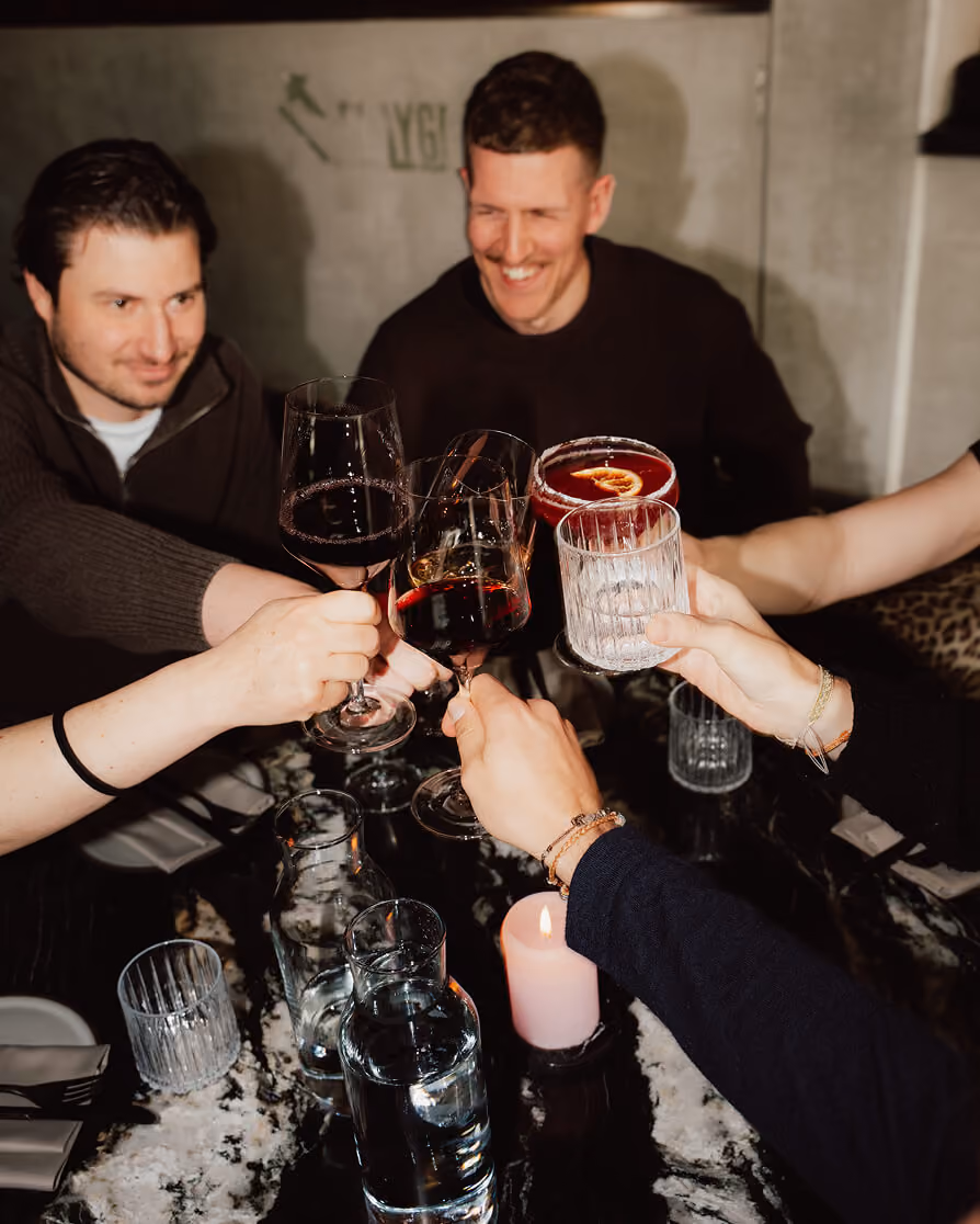 People clinking glasses with wine and cocktails over a dark marble table with a lit candle.
