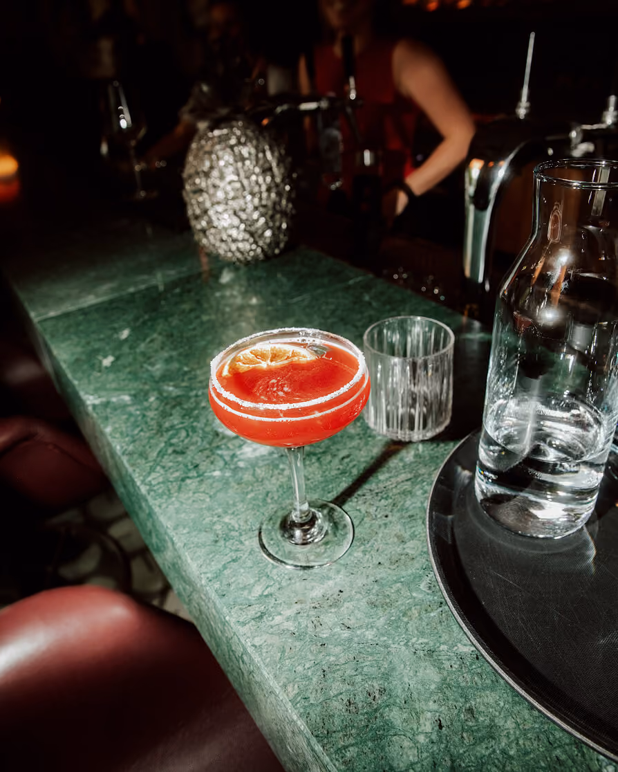 Red cocktail with salted rim and orange slice on a green marble bar counter in a dim setting.