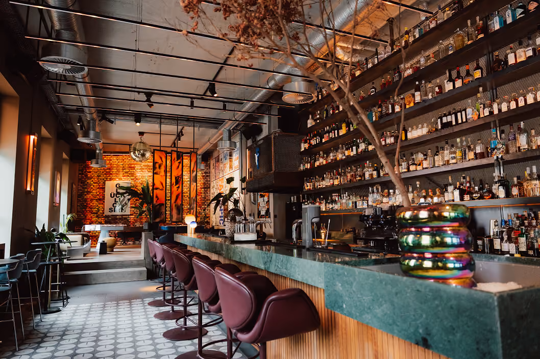 Modern bar interior with green marble counter, purple stools, extensive liquor shelves, and cozy seating area.