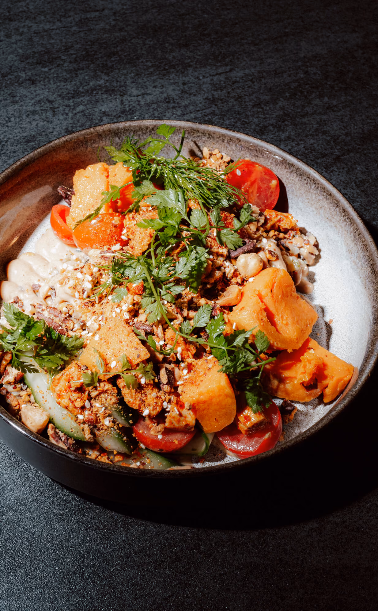 Bowl with chunks of orange sweet potato, cherry tomatoes, greens, and sprinkled seeds on sauce.