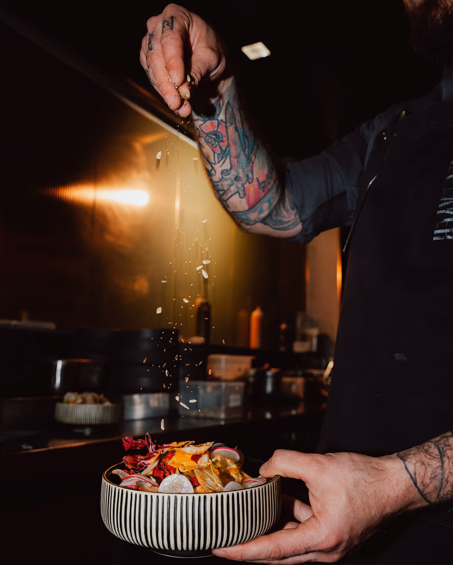 Tattooed chef sprinkling seeds on a colorful salad in a striped bowl in a dimly lit kitchen.
