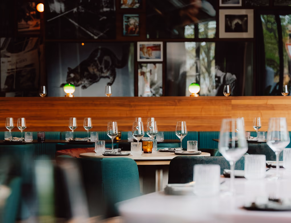 Modern restaurant interior with teal seats, round tables, glasses, and framed pictures on the wall.