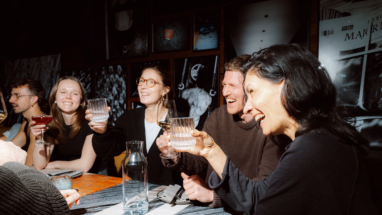 Group of five friends laughing and raising drinks while seated around a table at a restaurant.