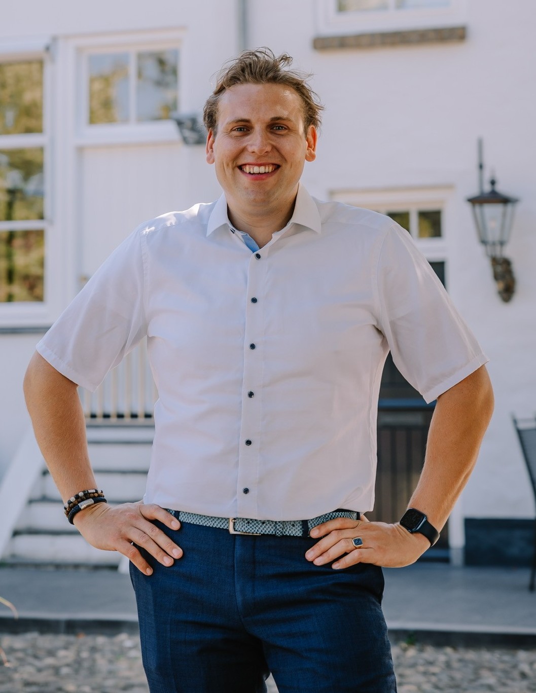 Sebastiaan, en man met kort blond haar, glimlachend, gekleed in een wit overhemd met korte mouwen en blauwe pantalon, staand met handen in de zij voor een witte woning.