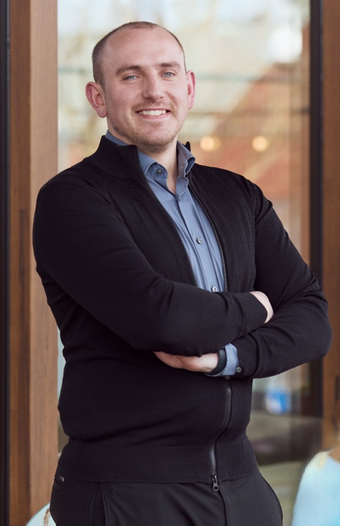 Yannick, een man met lichtblauw overhemd en zwarte trui staat met armen gekruist en lacht vriendelijk.