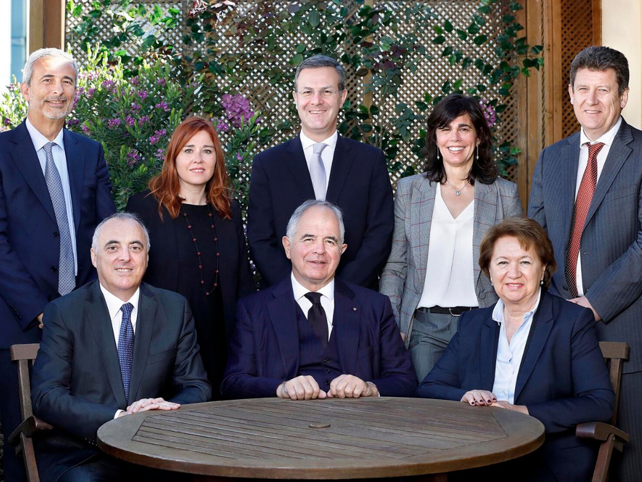 Een groep van acht volwassenen in formele kleding poseert buiten bij een ronde houten tafel met hedera en bloemen op de achtergrond.
