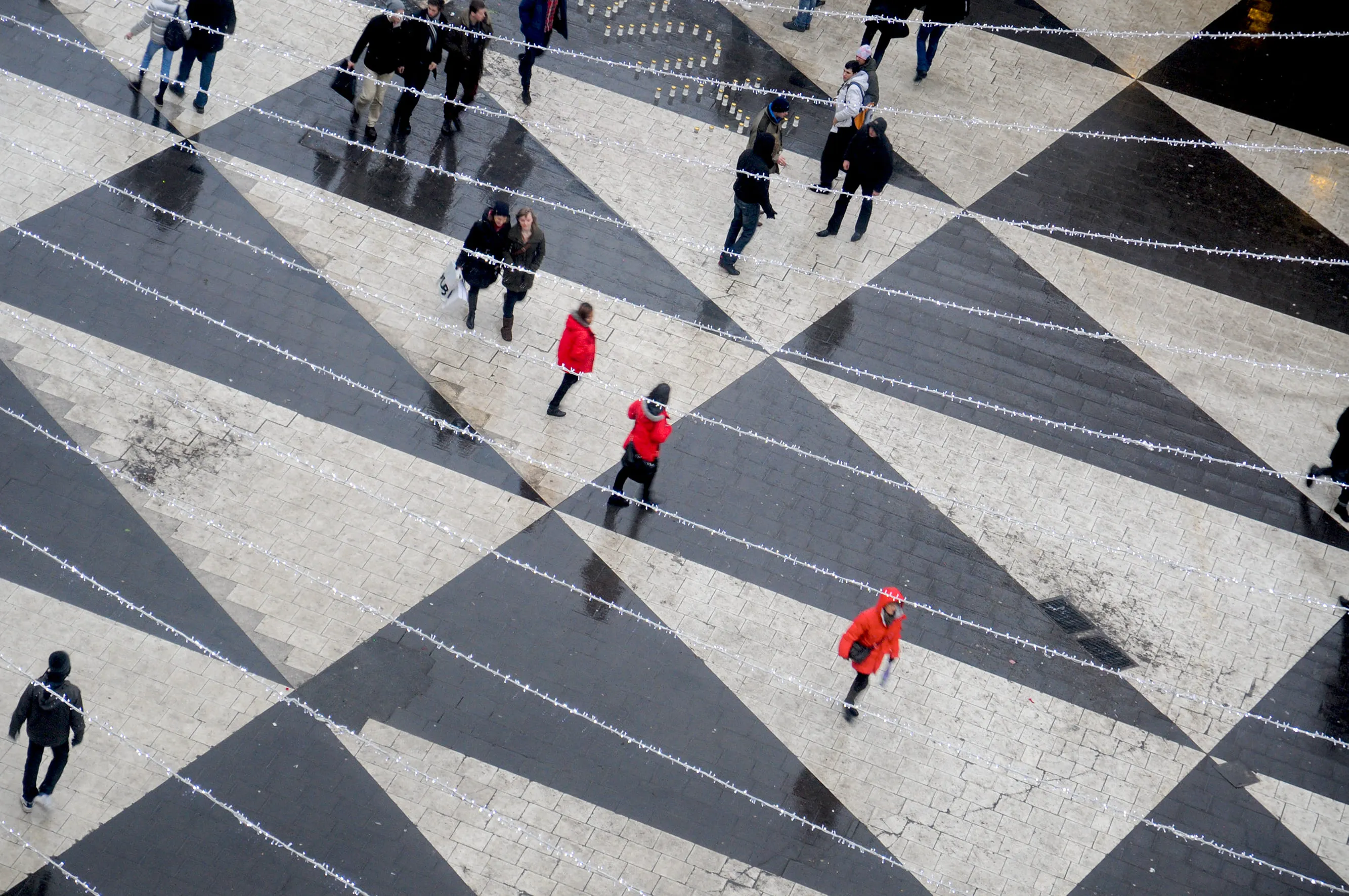 Place pavée avec un grand motif géométrique en noir et blanc, des personnes marchant avec des manteaux rouges, et des guirlandes lumineuses suspendues en travers.