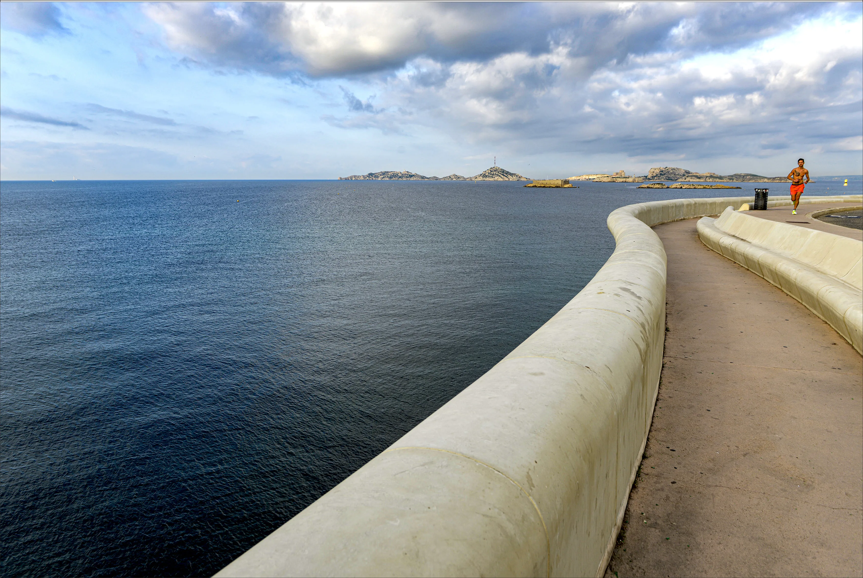 Un homme en short rouge court le long d'une promenade au bord de la mer avec des îles au loin sous un ciel nuageux.