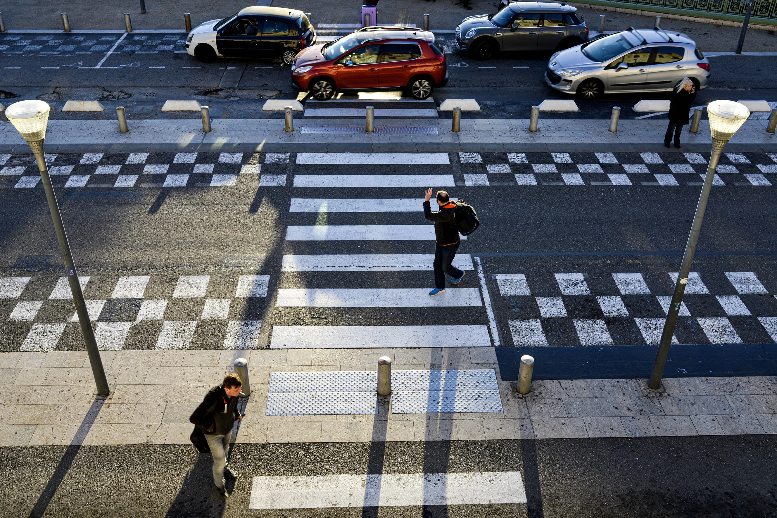 Vue aérienne d'un passage piéton avec trois personnes traversant une rue devant des voitures garées au soleil.