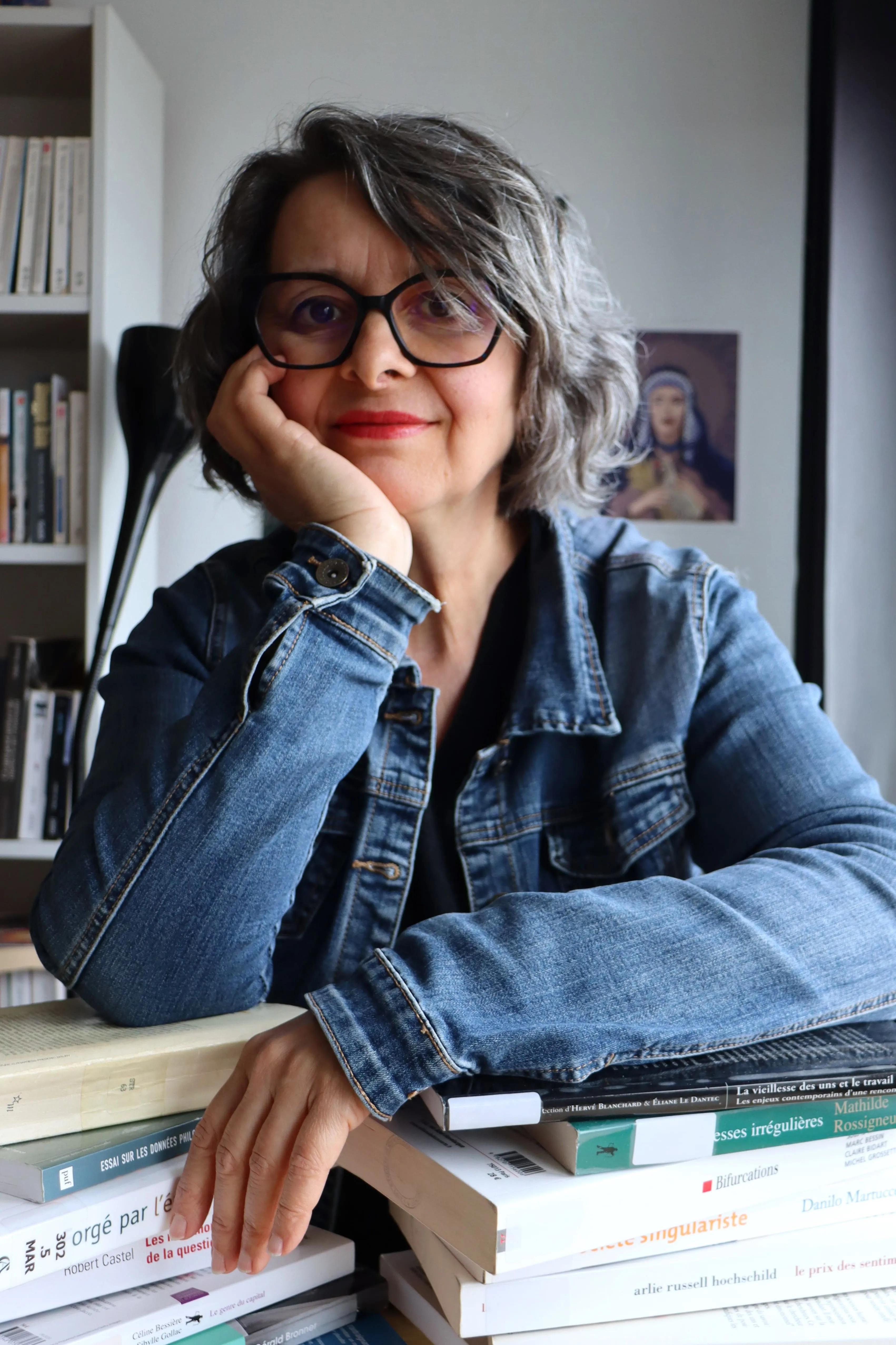 Christine Vallin Femme souriante aux cheveux gris courts et lunettes, appuyée sur une pile de livres, portant une veste en jean dans une pièce avec une bibliothèque en arrière-plan.
