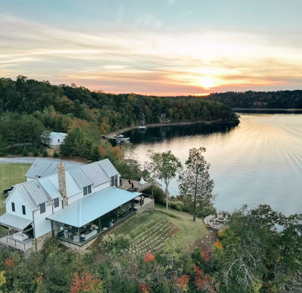 A large house with a metal roof overlooking a lake at sunset, surrounded by trees and greenery.