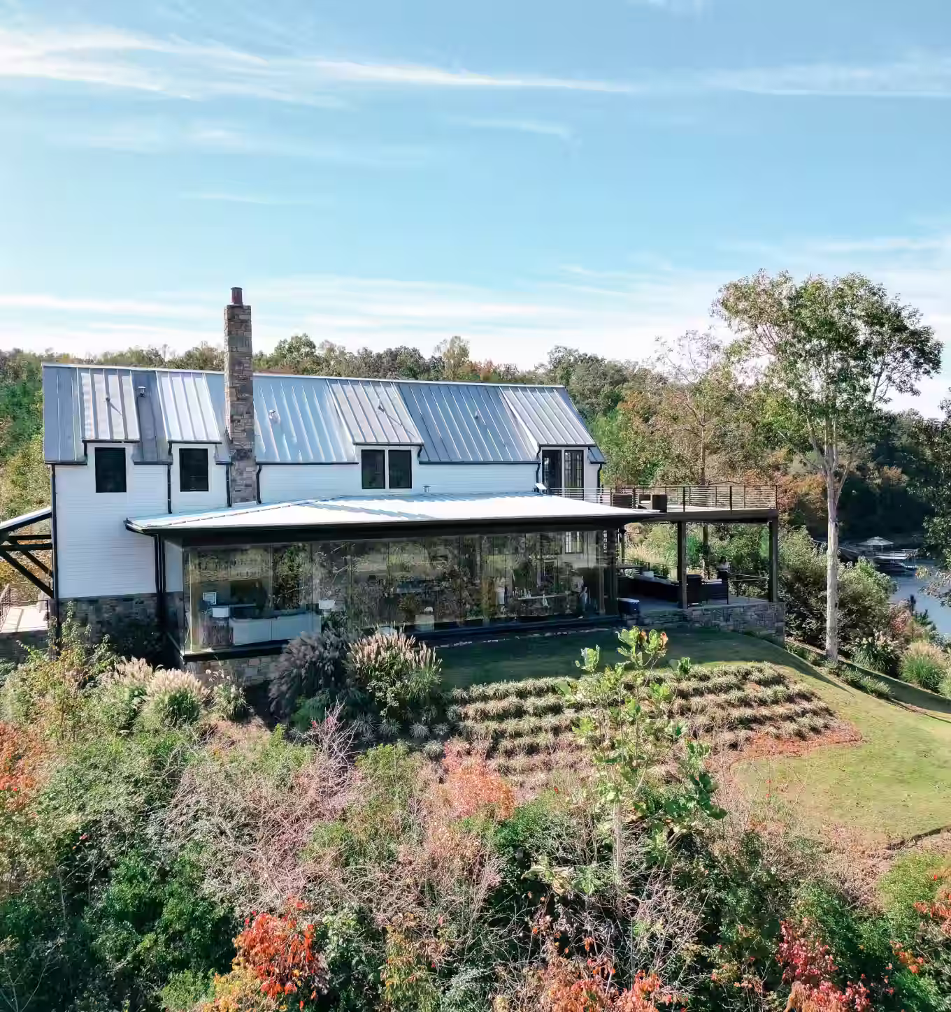 Modern two-story house with metal roof, large glass windows, surrounded by landscaped garden and trees near a body of water.