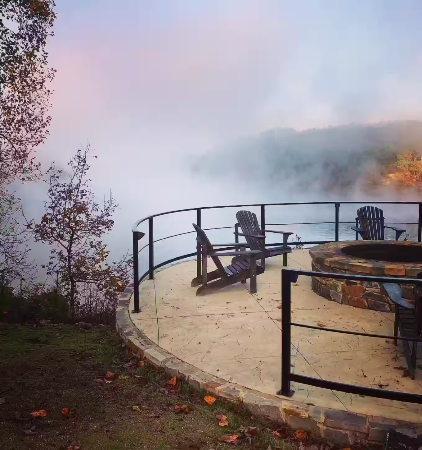Circular outdoor fire pit area with Adirondack chairs overlooks a misty lake surrounded by trees.