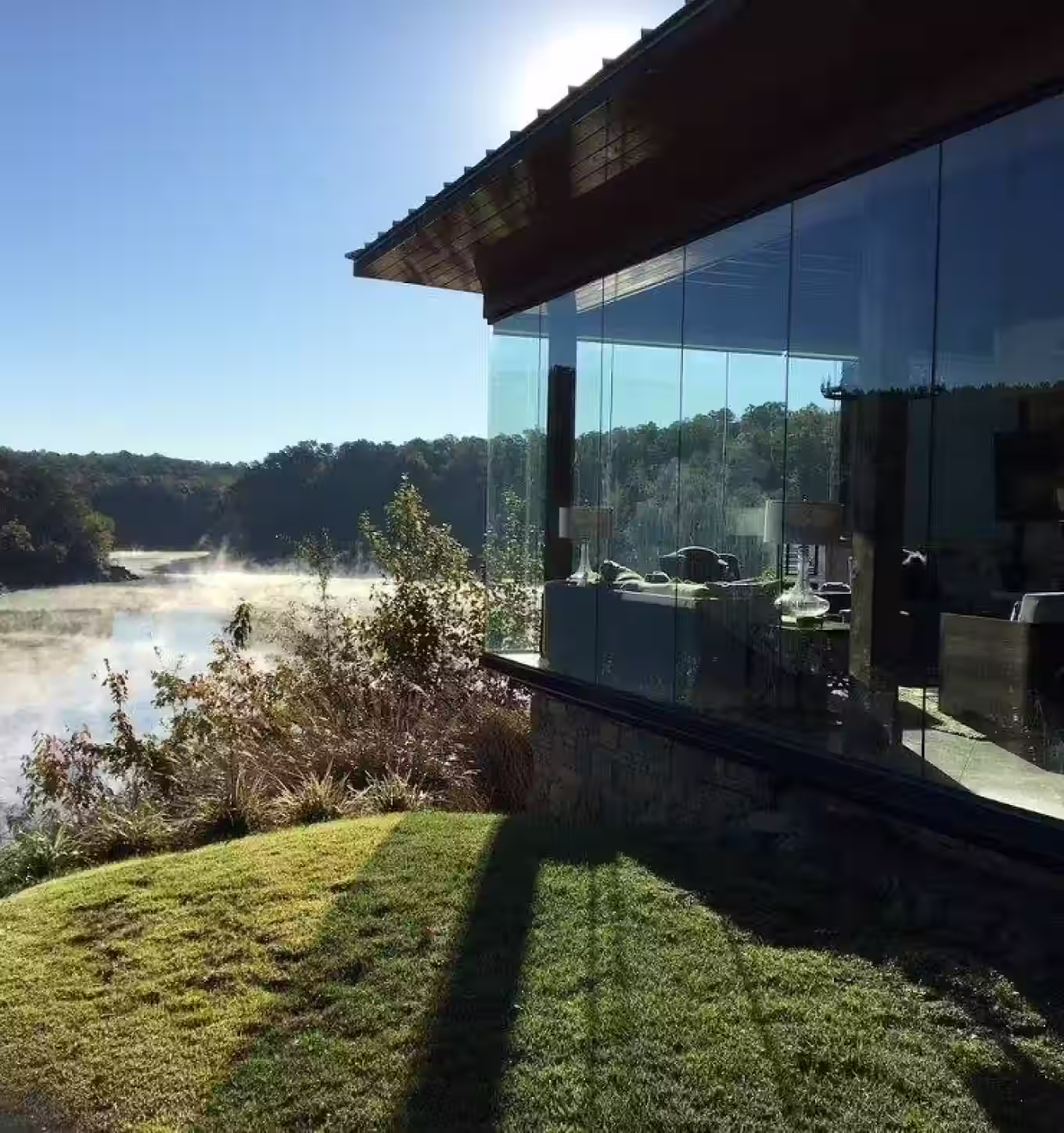 Modern glass-walled house overlooking a misty river surrounded by trees and greenery on a clear sunny day.